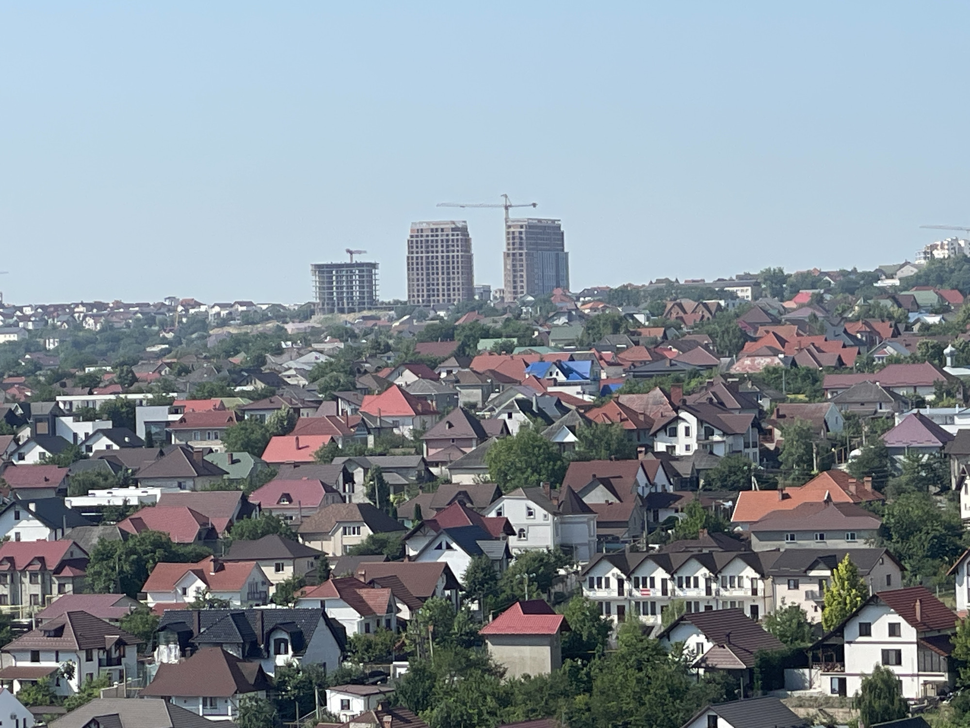 A wide, high-angle shot captures a dense residential area with numerous houses and some multi-story buildings under construction under a clear, light blue sky. The foreground is filled with a close-packed collection of houses featuring a variety of roof colors, including red, brown, and some grey, with many houses having pitched roofs. The houses are of different sizes and architectural styles, with some appearing to be two stories high with white or beige exteriors. Green trees are interspersed throughout the residential area, providing visual breaks. In the background, three tall, modern buildings are under construction, with visible cranes and scaffolding. These buildings are positioned on a slight rise and appear to be significantly taller than the surrounding houses. The overall impression is of a developing urban or suburban landscape.