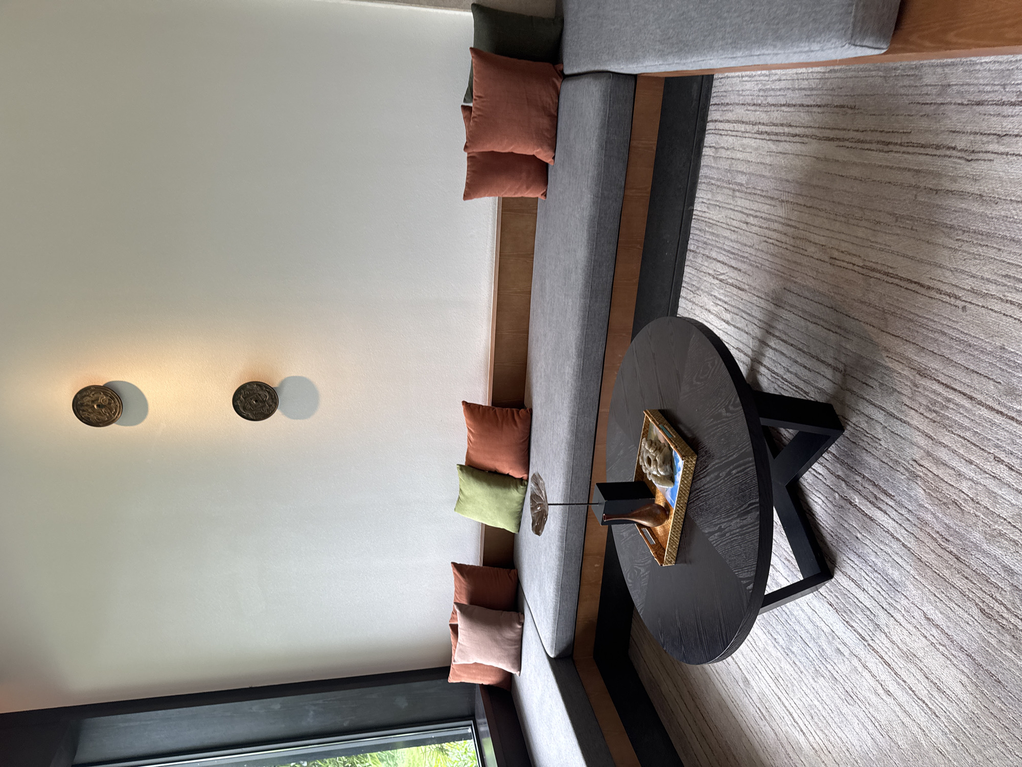 The image captures an interior lounge setting in what could be a hotel or upscale residential space. A sectional sofa occupies the center, adorned with multiple throw pillows in muted earthy tones, complementing the grey upholstery. The sofa is arranged in a corner configuration, hinting at a modern design aesthetic. A dark, round coffee table sits centrally, displaying a decorative object in a golden frame. The flooring is a light-colored carpet with a subtle linear pattern. On the adjacent wall, there are two circular decorative wall hangings. Light streams in from a window at the bottom of the frame, suggesting a garden or outdoor view. Given the lack of visible urban context, the assumption of Guilin, China, cannot be verified within the frame. The scene lacks human subjects and focuses on a staged, designed interior space.