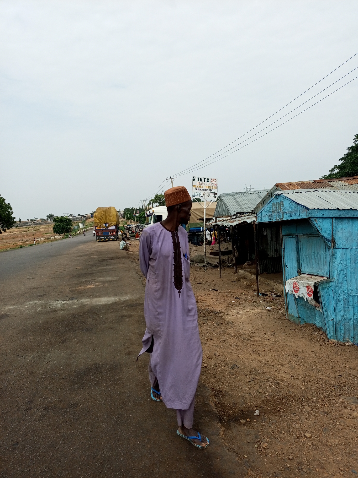 An adult male, dressed in a light purple traditional garment and a brown woven hat, stands facing right and looking forward, his body angled slightly away from the camera. He is wearing blue flip-flops. The man is positioned on the right side of a dusty road in Gadao, Nigeria. To his right, a small, vibrant blue corrugated metal structure, likely a shop or kiosk, is visible, with some signs and papers affixed to its front. Behind the blue structure, more corrugated metal buildings with rusty roofs are lined up. A signpost with "NURTW HEAVY TRUCK NERXMILE THREE GUNDE GUNDE STATE" is also present. To the left of the man, the road stretches into the distance, with a yellow-covered truck visible in the middle distance, along with other vehicles and pedestrians. The overall environment appears to be a roadside market or settlement under an overcast sky.
