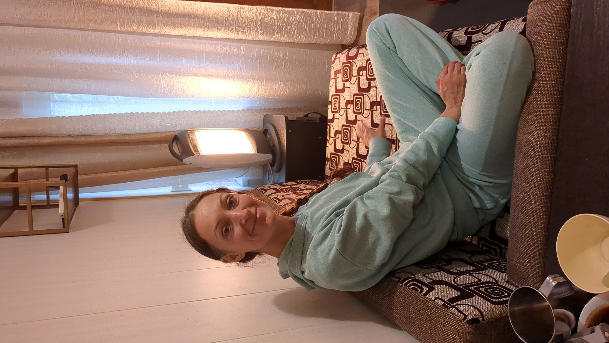 An adult woman is pictured reclining on a patterned sofa in what appears to be a cozy indoor setting, possibly a living room, in Gelendzhik, Russia. She is smiling gently at the camera, conveying a relaxed and content emotion. Her dark hair is pulled back, revealing her face. She is dressed in a comfortable light blue (or mint green) hooded tracksuit, with her hands visible resting on her knee and near her side.
The sofa she is on features a brown and cream geometric pattern with swirls and squares, and the visible armrest is dark brown and textured. In the background, to her left, a rectangular heater or lamp emits a bright, warm light, suggesting it's either evening or a dim environment, though some natural light filters through a window behind light-colored, patterned curtains or blinds. A small, light-wood shelving unit is visible further to the left. The walls are white. In the bottom right foreground, out-of-focus household items like a light-colored plastic cup and a silver metal cup are visible on a surface next to the sofa. The overall atmosphere is one of comfort and leisure. No visible text is present.