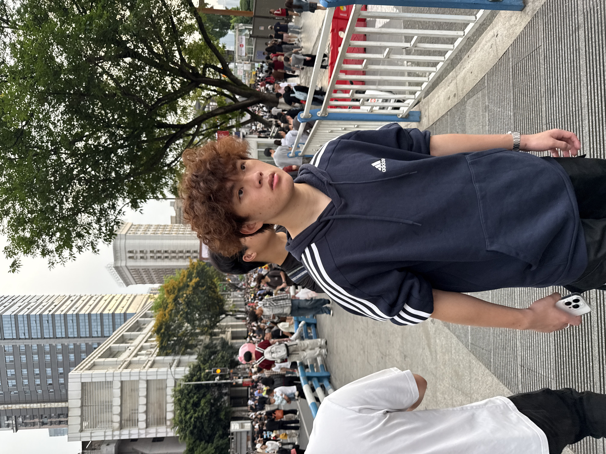 A young man with curly brown hair stands on a sidewalk in the Yuzhong District of China, wearing a navy blue Adidas hoodie and black pants. He holds a white smartphone in his right hand. Behind him, a large crowd of people is visible in the distance. The background includes trees, tall buildings, and metal railings. Another person wearing a white shirt is partially visible in the lower portion of the image. The sidewalk surface is textured with lines.