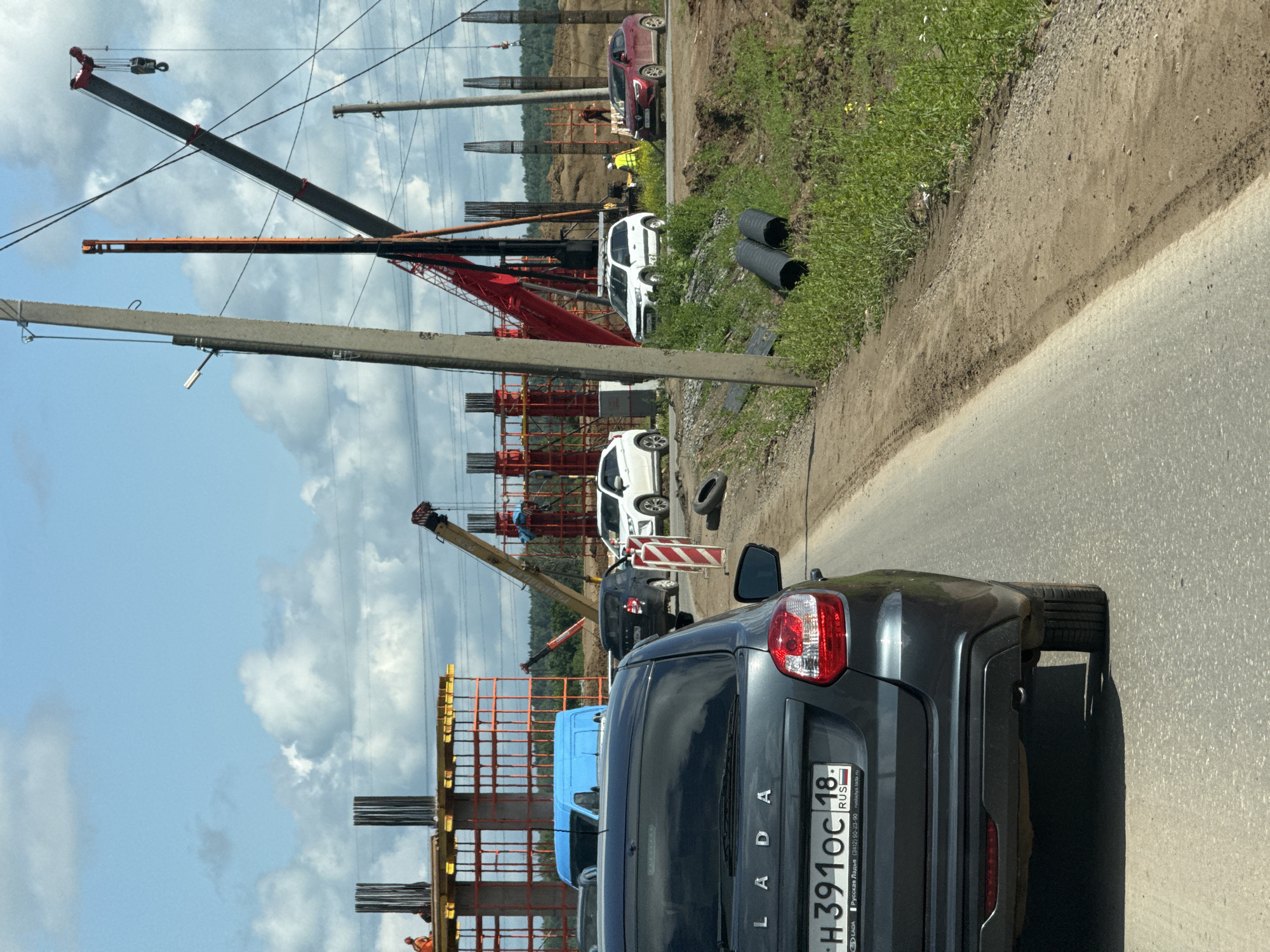 The image depicts a construction site on a sunny day with a cloudy sky. In the foreground, the rear of a dark gray Lada sedan is visible, with its license plate reading "H391OC 18 RUS". The car is on a paved road, with a dirt and grass shoulder to its right.
The construction site features several large concrete pillars under construction, with rebar exposed at their tops. Orange scaffolding is erected around some of these pillars. Two large cranes are present, one red and one yellow, actively involved in the construction process. Several workers in orange vests and hard hats are visible on the site. Other vehicles, including a blue van, a black sedan, and two white SUVs, are parked or driving on a road within the construction area. In the background, a wooded area is visible. The scene suggests ongoing infrastructure development. The location is identified as Kabanikha, Russia.
