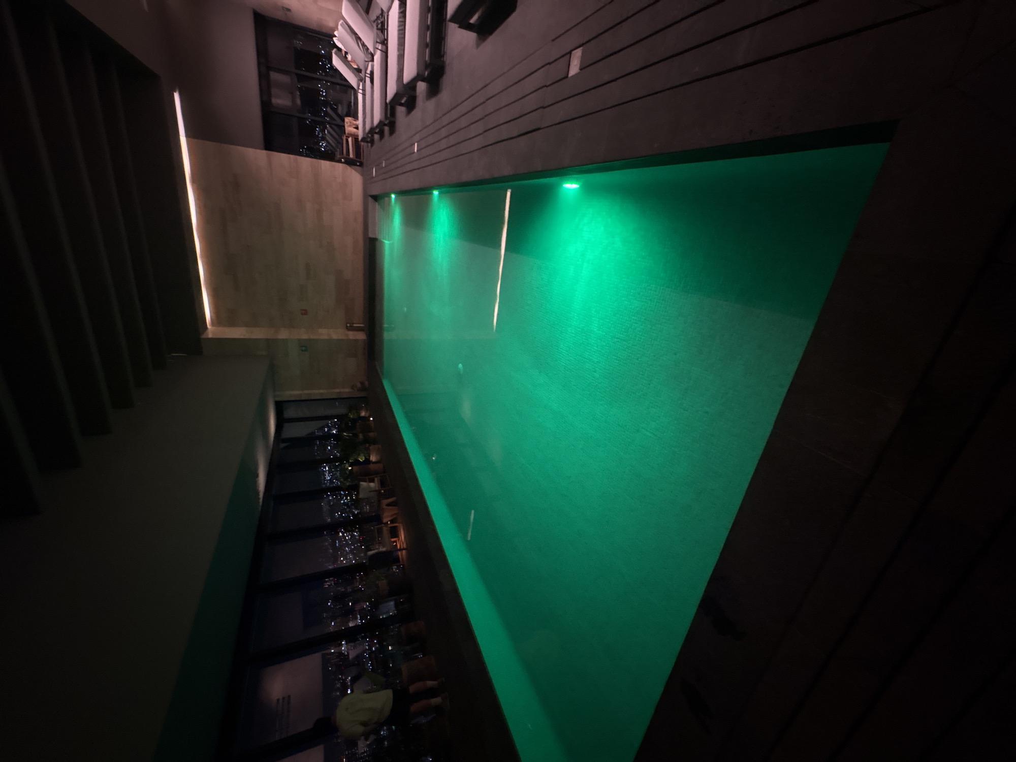 The image shows a rooftop swimming pool in Jesús del Monte, Mexico. The pool is lit with green lights and appears to have an infinity edge. A glass-walled building, presumably a restaurant or bar, is adjacent to the pool. The pool offers a panoramic view of the cityscape below, with city lights visible through the glass wall. The area around the pool is dimly lit.