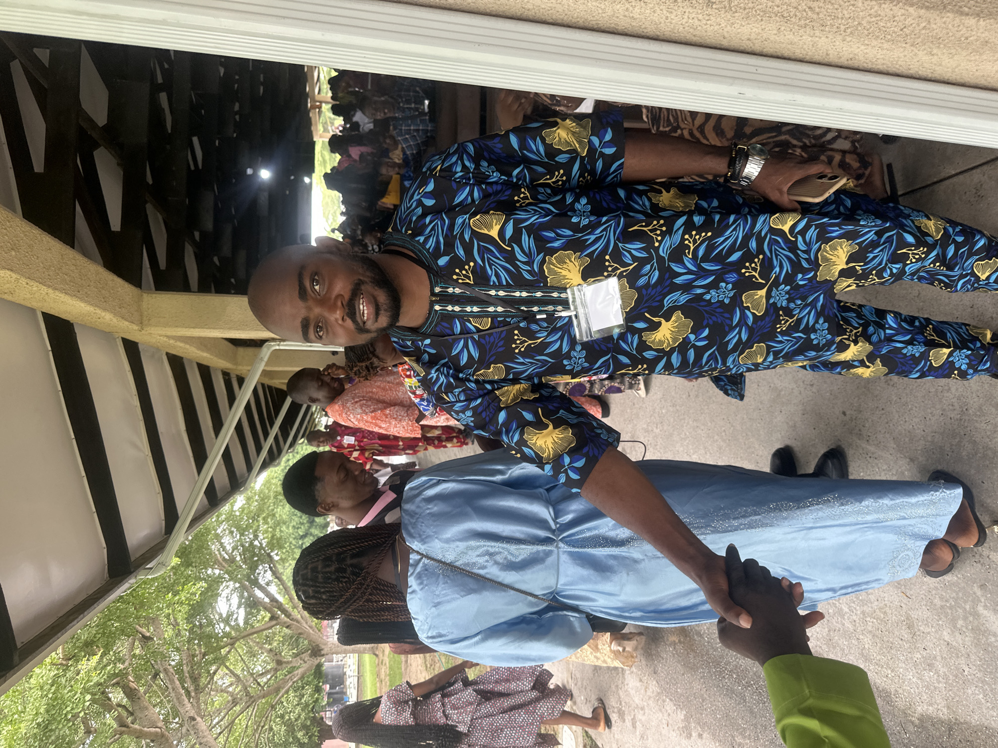 In Itoga, Nigeria, a man in a patterned blue and yellow dashiki shakes hands with another person whose arm and hand are visible on the left, wearing a lime green garment. The man in the dashiki is facing the camera, smiling, and appears to be wearing a lanyard with a badge around his neck. He is holding a smartphone in his right hand. Behind him, a group of people are gathered under a covered walkway with a visible roof structure. To the left of the man, a woman in a light blue dress is also visible from the back, her hair in braids. The environment suggests an outdoor gathering, possibly a community event or a religious service, with a concrete walkway and trees visible in the background.
