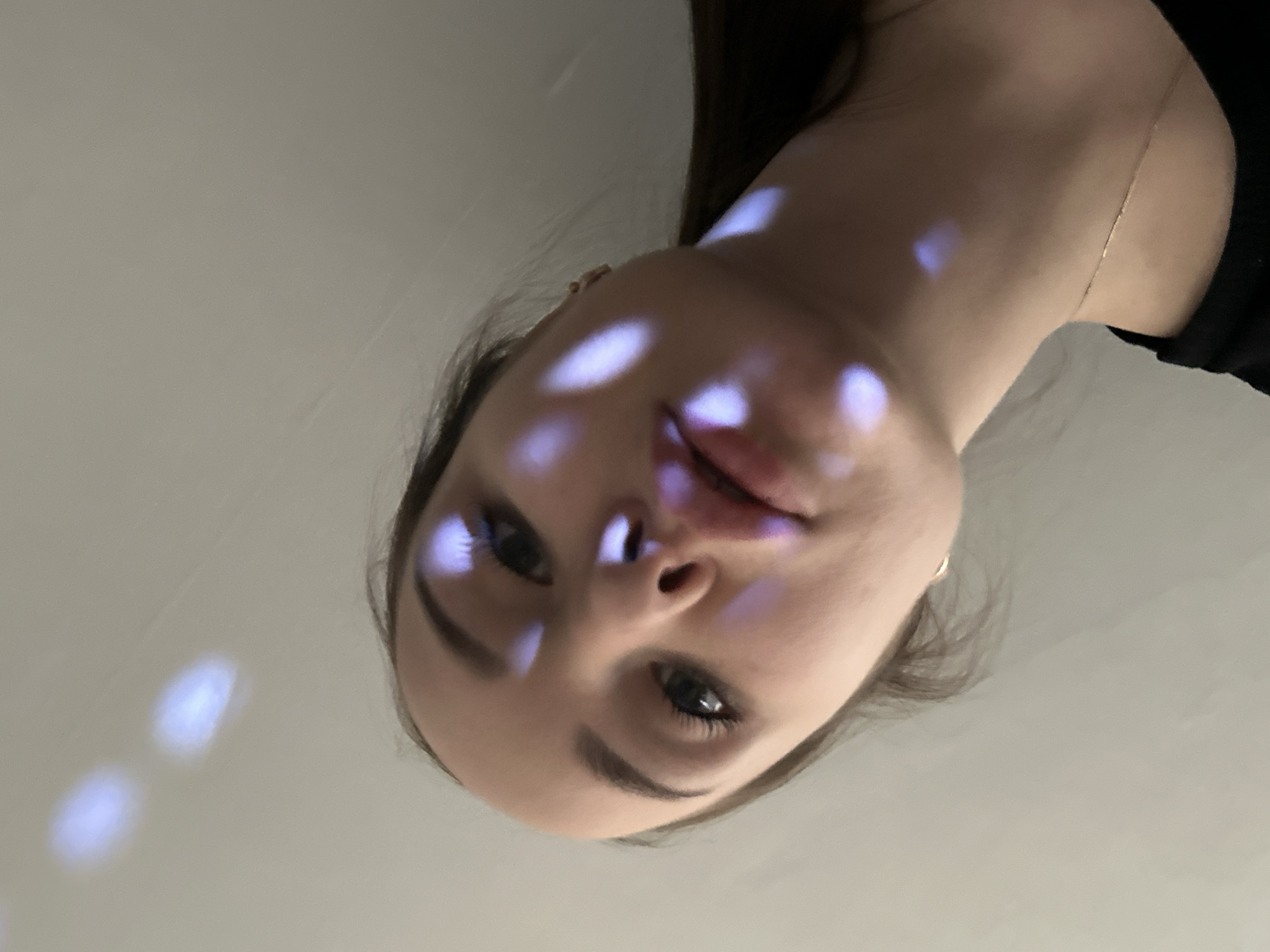 An artistic, low-angle portrait captures the face of a young woman, likely taken indoors in Chaikovsky, Russia. Her head is tilted, and she gazes directly at the viewer, with a neutral to slightly contemplative expression. Her dark hair frames her face, and a subtle metallic chain is visible near her neck. The most distinctive feature is the unique lighting: several diffused, blue-ish white spots of light are cast across her forehead, cheeks, and lips, as well as on the plain, light-colored background. This patterned illumination suggests a specific light source, such as a disco ball or projector, lending an ethereal quality to the image. Her facial features are clearly visible, including her eyelashes, eyebrows, and natural lips. The background is a minimalist, uniform light grey/off-white surface, providing a stark contrast to the subject. No specific activity or event is explicitly shown beyond the act of posing for the photograph. There is no visible text in the image.