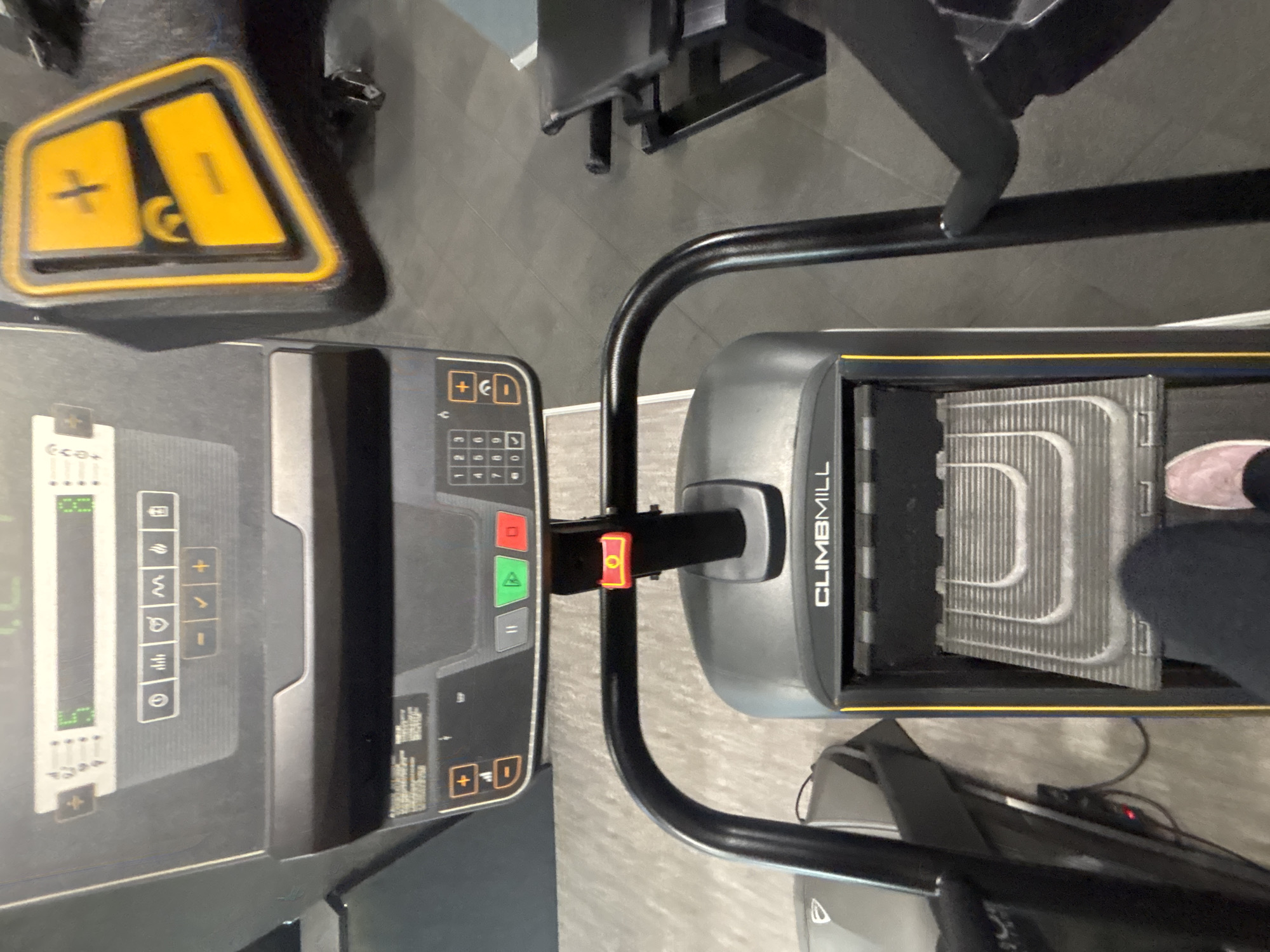 The image shows a top-down view of a gym setting, featuring fitness equipment. Dominating the frame are two pieces of equipment: a treadmill with a black and gray control panel and a stair climber branded "CLIMBMILL." A person wearing dark pants and light pink shoes is standing on the stair climber. A piece of black exercise equipment with a yellow button is next to the treadmill. The floor is a light gray color. There is nothing to indicate this photo was taken in Varna, Bulgaria.