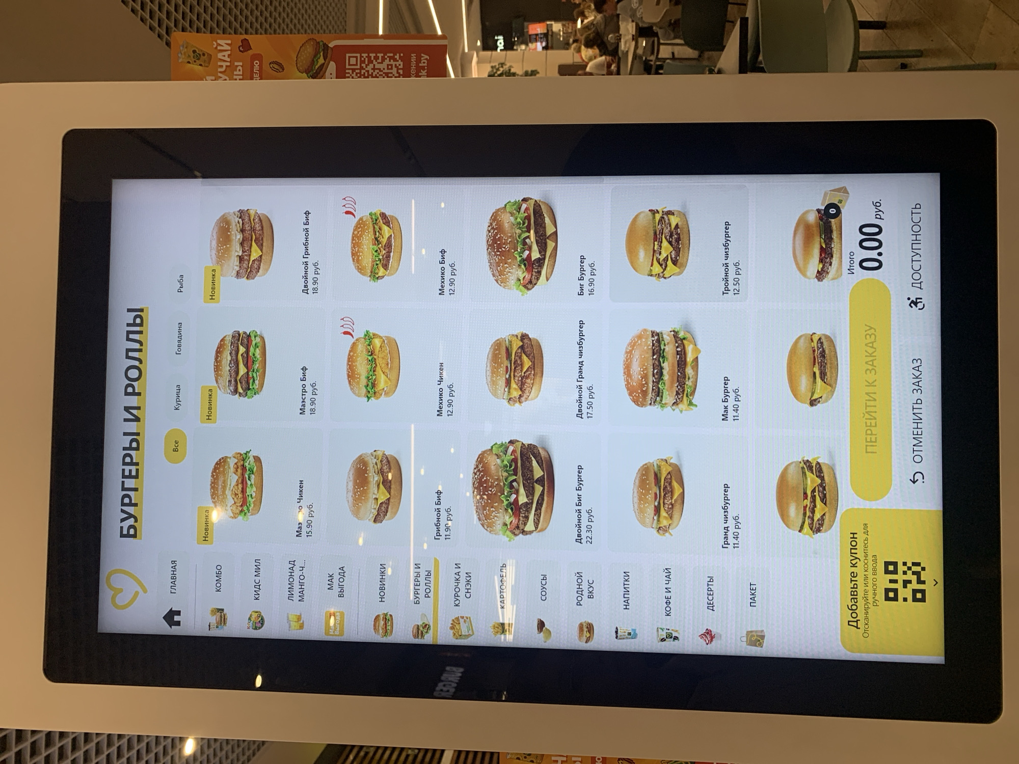 This is a photograph of a digital menu screen inside a McDonald's restaurant. The screen displays a selection of burgers and rolls, with prices listed in Russian rubles. The menu is divided into categories such as "Burgers and Rolls," "Combos," "Kids Meal," and "Drinks." Some items are marked as "New" or "Special Offer." At the bottom of the screen, there is an option to scan a coupon or enter a code.
The restaurant environment appears to be clean and modern, with bright lighting. The background shows other patrons seated at tables and a chair. The overall impression is that of a typical fast-food restaurant.
Based on the context provided by the user, this McDonald's is located in the city of Hrodna, Belarus.