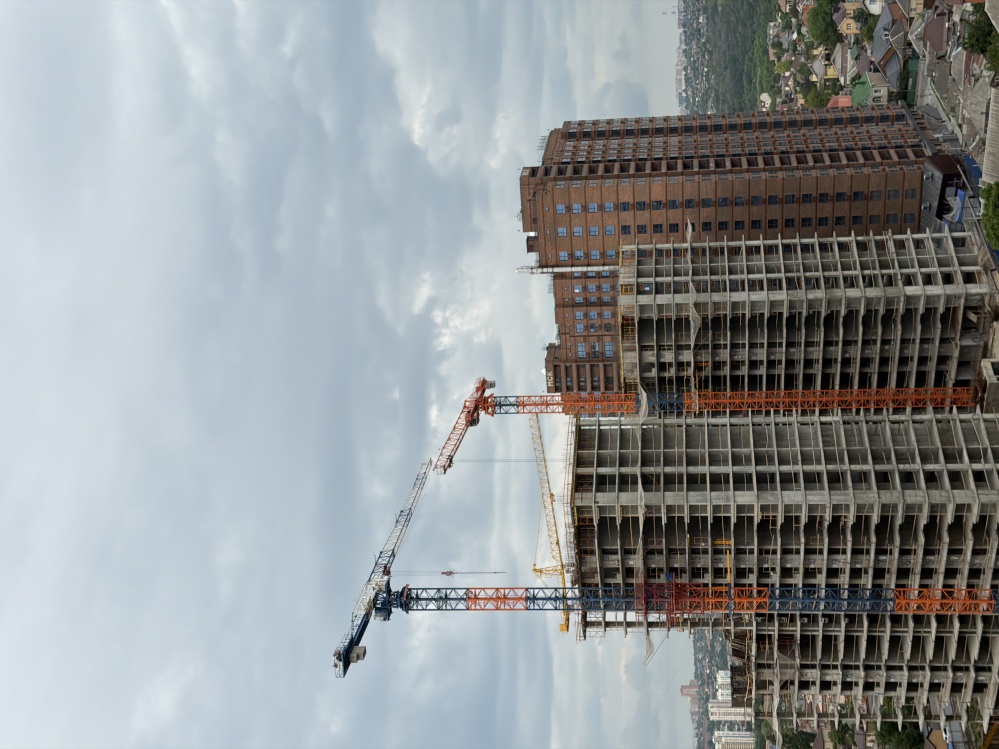 The image shows a construction site in the city of Chaltyr', Russia. A multi-story brown building stands next to a concrete skeleton of another high-rise under construction. Several tower cranes are positioned around the construction site, with one painted in blue, orange, and white, and another painted yellow. The sky is overcast. In the background, a residential area is visible.