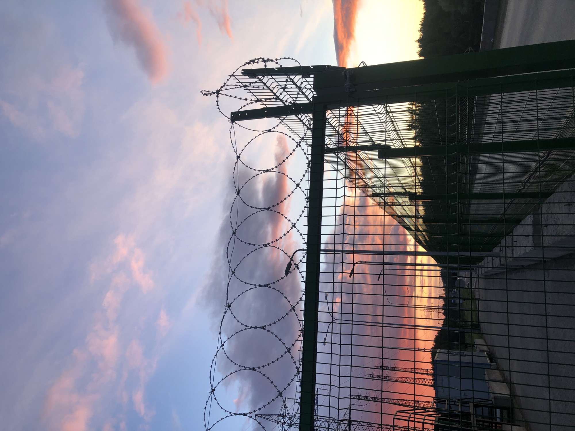 A green chain-link fence topped with coiled barbed wire dominates the frame. The scene beyond the fence includes a street lined with utility poles and structures, silhouetted against a vividly colorful sky at sunset. The sky's colors are reflected in the gaps of the chain-link. The fence appears to be located in an industrial or utility area, given the presence of the poles and buildings. The scene is potentially located in Noviy, Russia, per the provided instructions. There are no people visible.