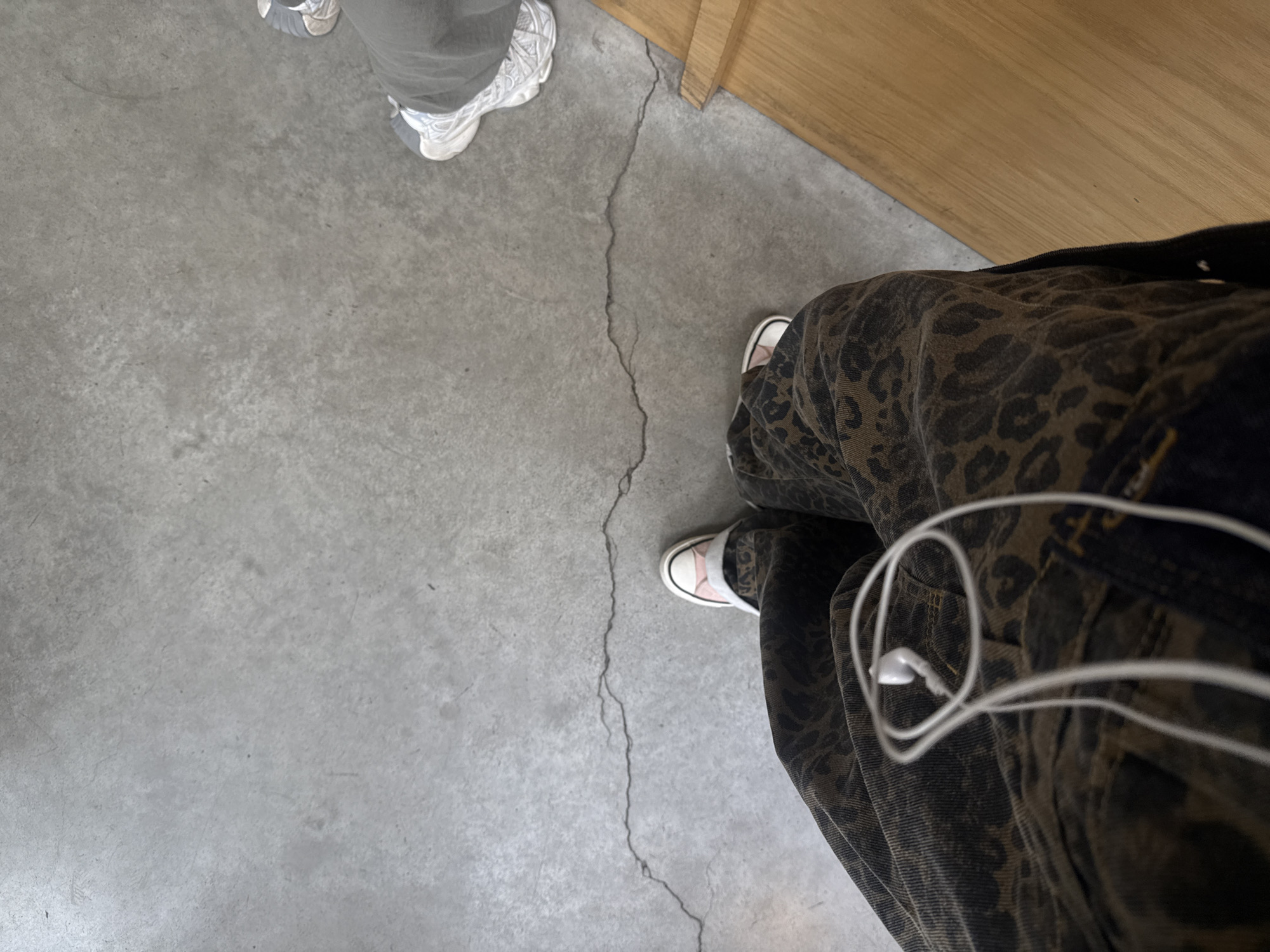 The image shows a view looking down at two pairs of feet standing on a concrete floor. The feet in the upper left are wearing grey sneakers, while the feet in the lower right are wearing white sneakers with a light pink toe cap. The legs of the person in the foreground are clad in pants with a dark leopard print pattern. White earbud wires are visible next to their pants, seemingly attached to a device in the pocket. A light-colored wooden surface is present at the top of the frame. A crack runs through the concrete floor. The location is not identifiable as Yerevan, Armenia, based on the visual information provided.