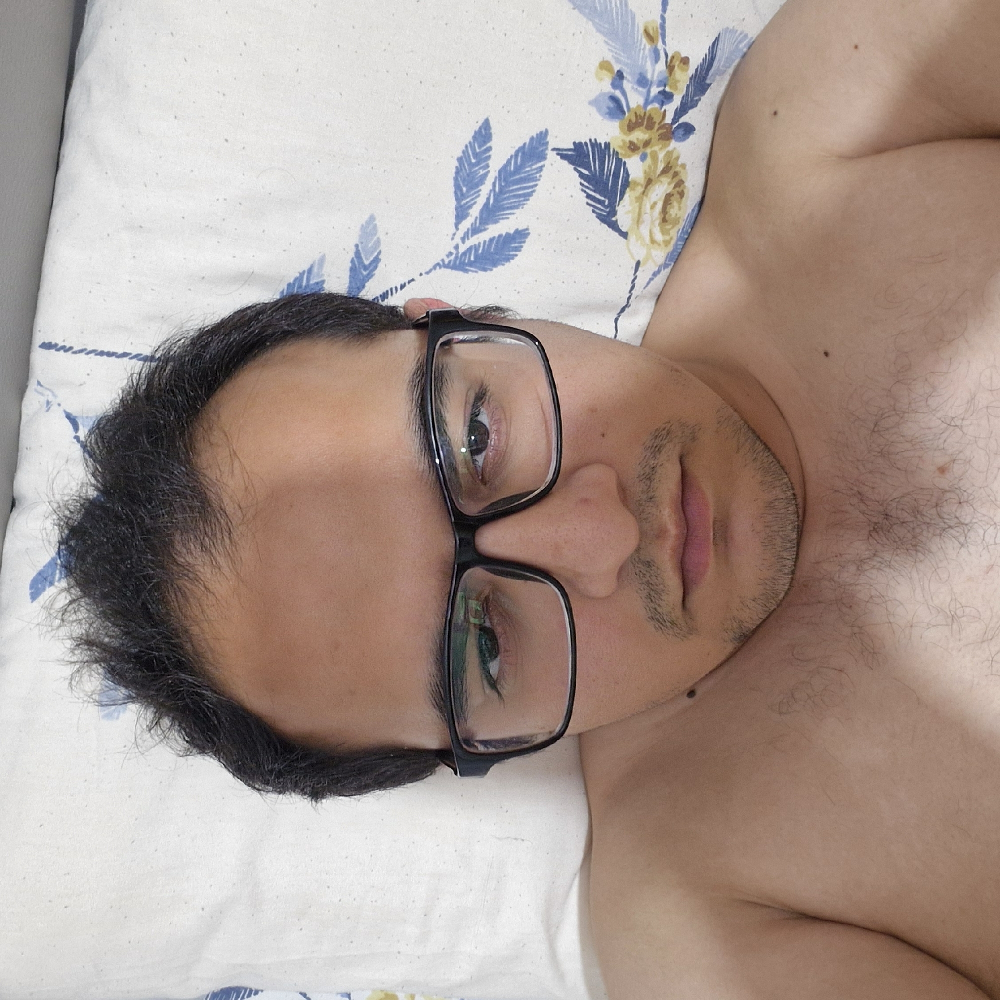 A close-up, overhead shot captures a person lying on a floral-patterned pillow, facing upwards. The person, wearing black-rimmed glasses, has dark, somewhat tousled hair and is shirtless, revealing chest hair. Their head is slightly tilted, and they appear to be looking directly at the camera. The lighting casts subtle shadows on their face. The background consists of the pillow and a light gray surface, possibly a bed. The specific location in Küçükçekmece, Turkey, is not discernible from the visual elements in the image.