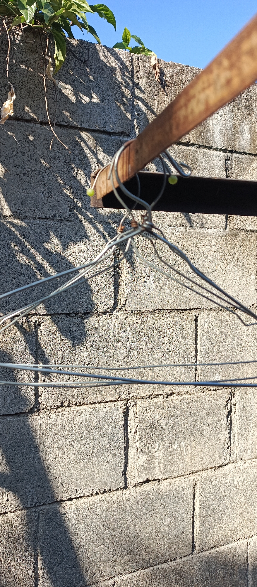 The image shows a weathered, rusty metal bar secured to a concrete block wall. Wire hangers are attached to the bar, creating a complex, tangled arrangement. Green beads are present on the wire. The wall is textured and shows signs of age. Overhanging the wall are green leaves and dry twigs, casting shadows on the wall. The sky is a clear blue. This scene is located in Jatisari, Indonesia.