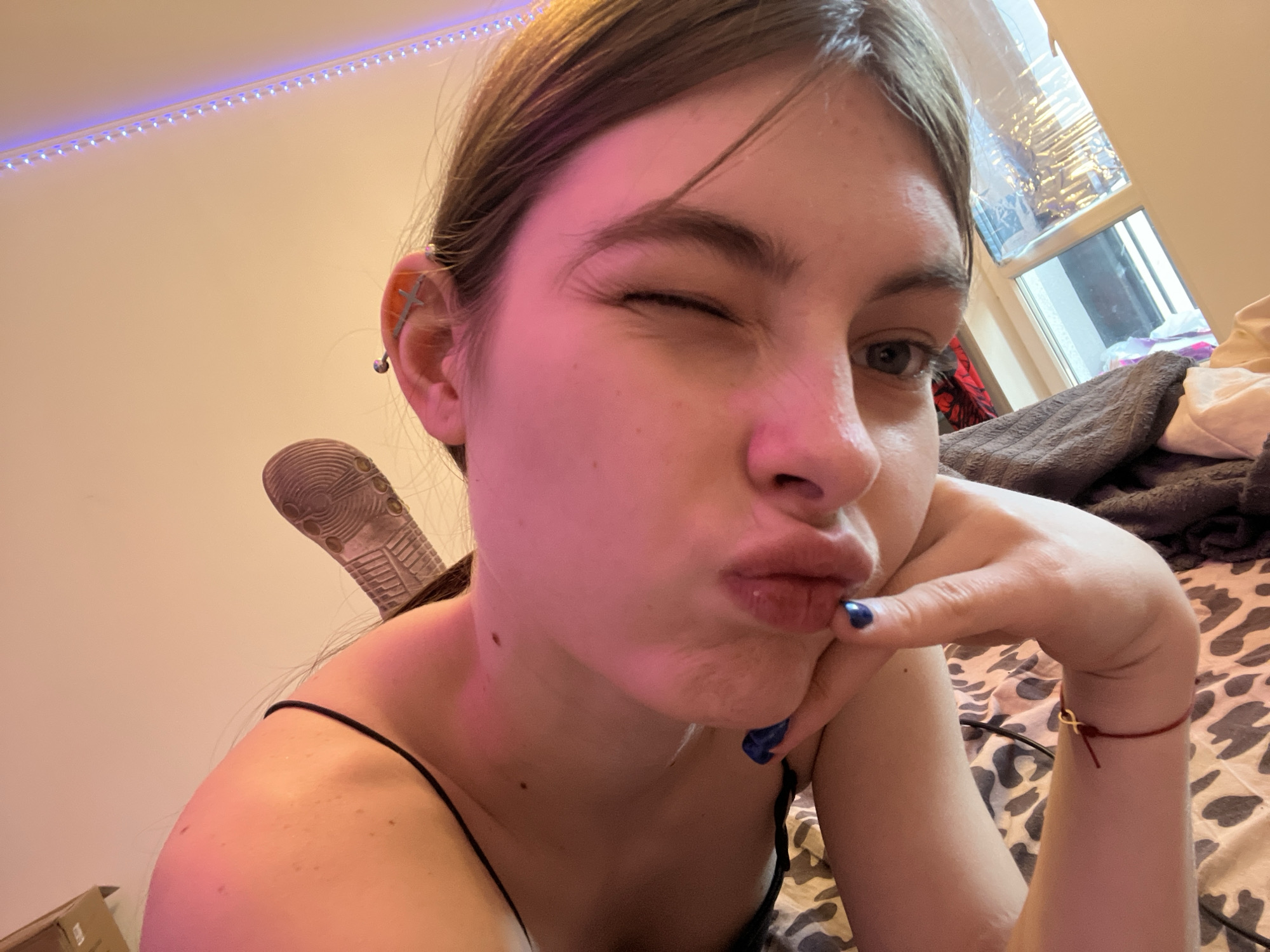 A close-up selfie shows a young woman with fair skin winking her left eye. Her lips are pursed in a pout. She has brown hair pulled back, revealing a pierced ear with multiple earrings. Her right hand is resting on her chin; blue nail polish is visible on her fingernails. A red string bracelet is around her wrist. The background includes a wall with a string of LED lights, a window with light filtering through blinds, and a bed covered in patterned bedding and a grey blanket. A sandal is near the woman's left shoulder. There is no evidence to support the location being Saint Petersburg, Russia.