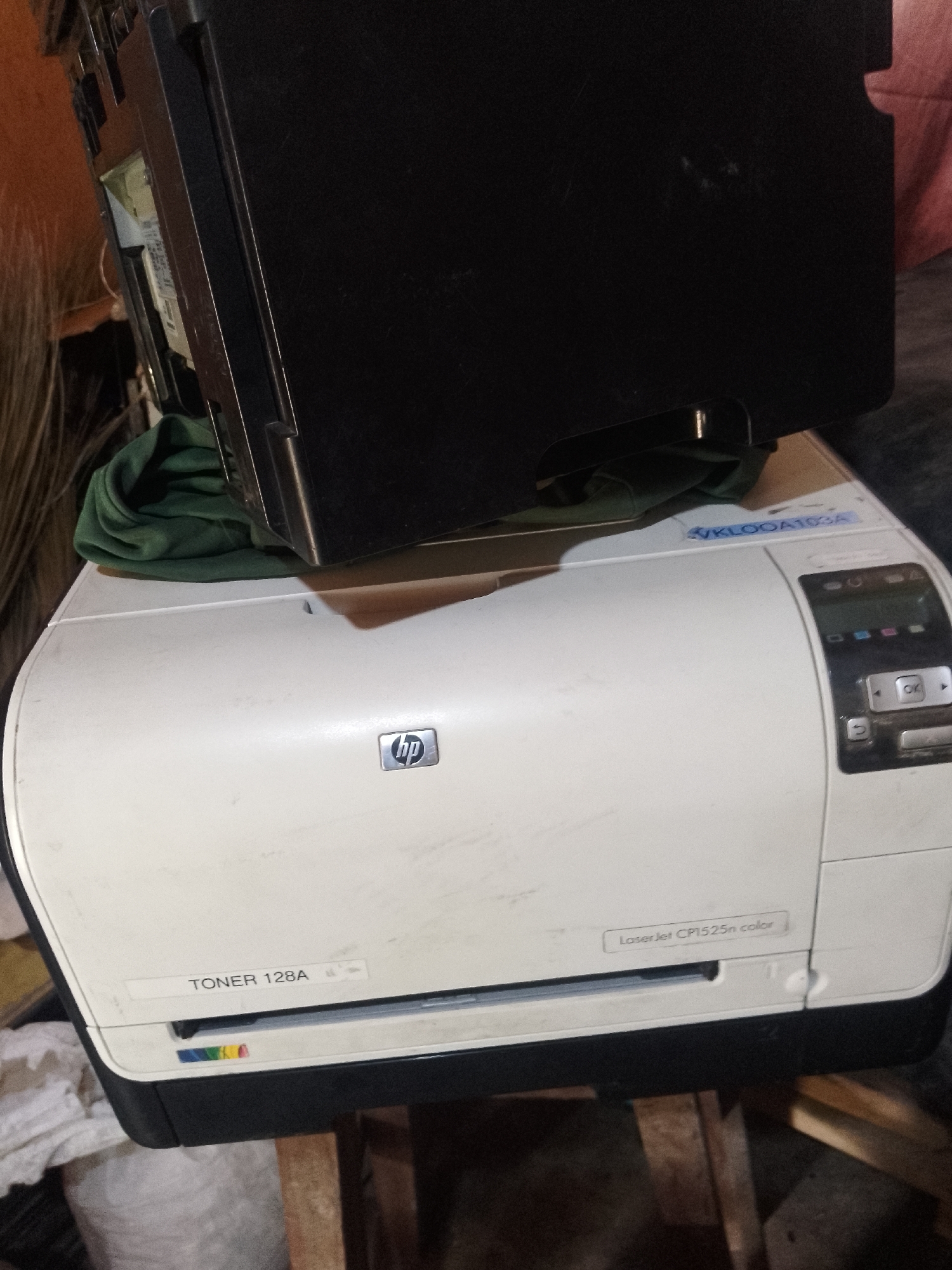 The image shows a white HP LaserJet CP1525n color printer in a dimly lit indoor environment. The printer is positioned on a wooden surface and is partially covered by a black object, possibly another part of a printer or scanner. A green cloth is draped over the top of the printer. A label on the front of the printer reads "TONER 128A" and another label on the top right indicates "LaserJet CP1525n color". The printer appears to be used and has some dust on its surface. The background is out of focus, but suggests an indoor setting with some clutter. There are no people visible in the image. The image does not provide any information about the city of Serti, Nigeria.