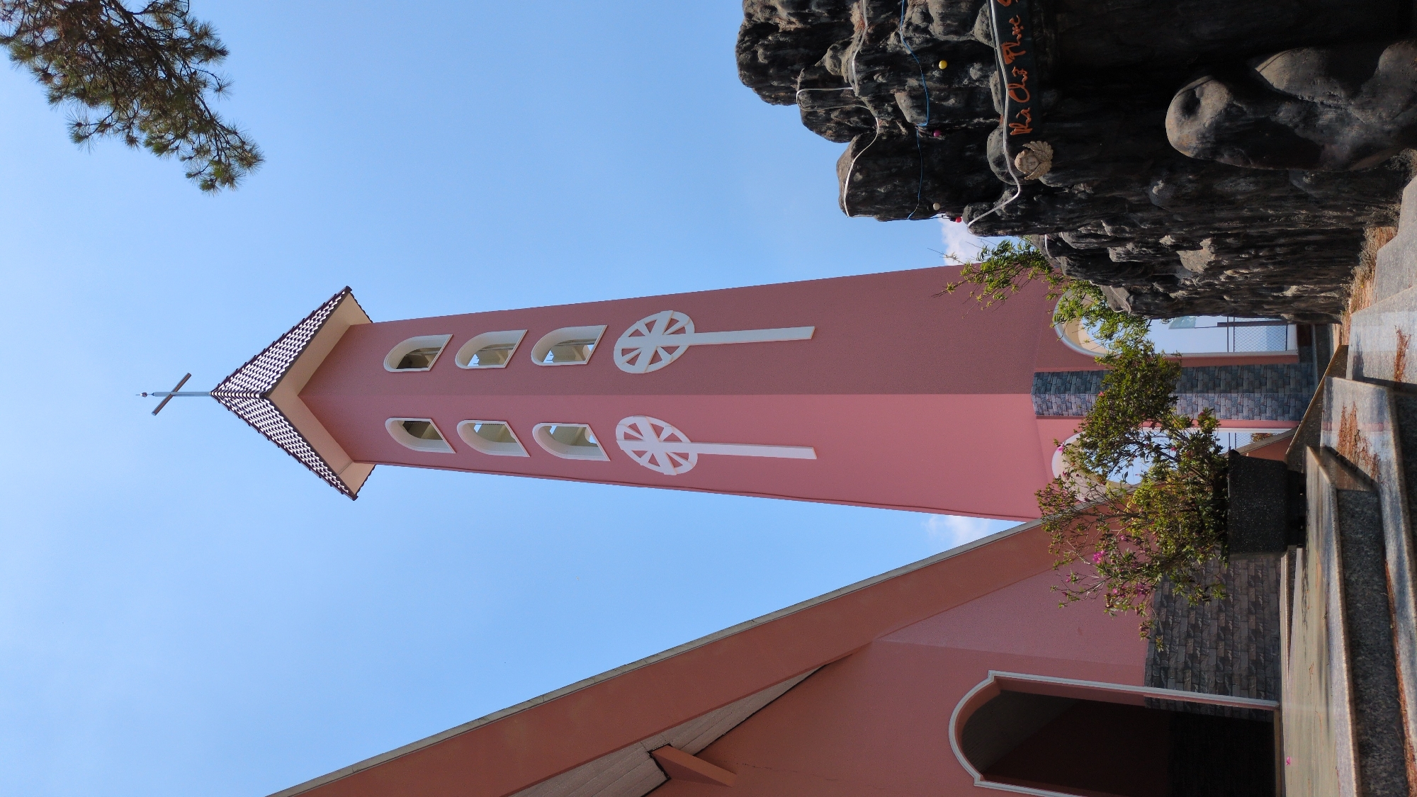 A low-angle shot of the bell tower of the Domaine de Marie church in Đa Thọ, Vietnam. The tower is pink with white accents, with a cross at its top. A series of arched windows are visible along the tower, and white decorative wheel motifs are also present. The blue sky is visible in the background, along with tree branches in the upper left corner. The base of the church is visible, along with a decorative rock feature and stairs to the right.