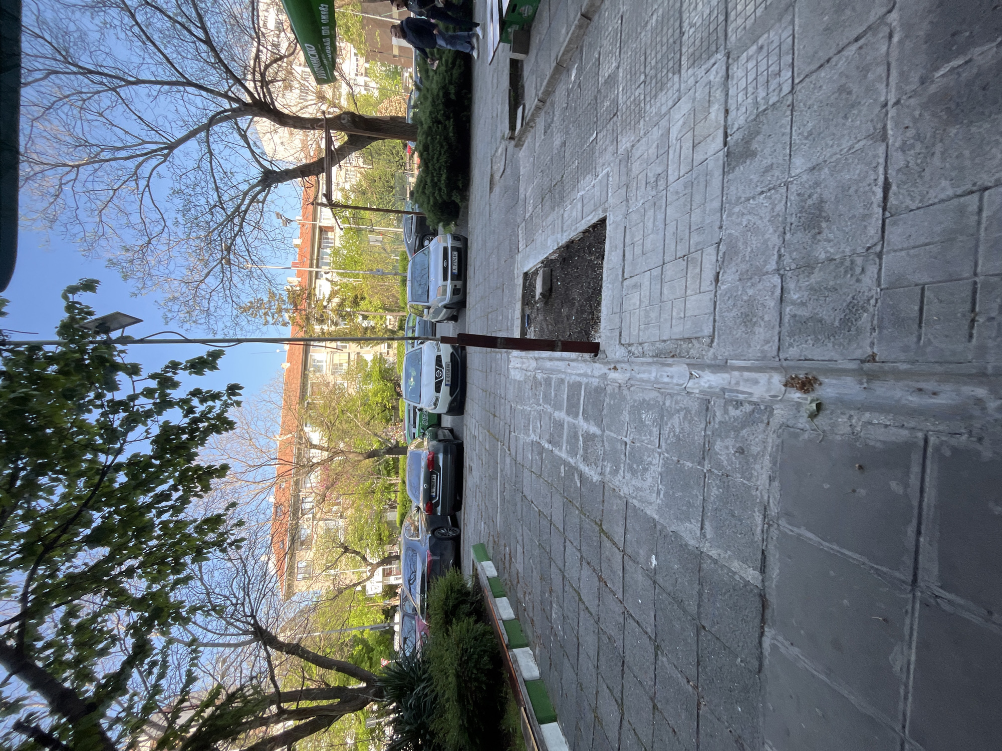 The image depicts a street scene in Burgas, Bulgaria. A row of parked cars lines the left side of the frame, next to a sidewalk paved with gray rectangular stones. A narrow strip of greenery, including bushes and trees, separates the sidewalk from the cars. The curb is painted with alternating white and green stripes. On the right side of the sidewalk, there is a rectangular patch of bare dirt. The buildings are visible in the background, primarily apartment buildings. People are walking on the sidewalk. Overhead, branches of trees reach towards a clear blue sky.