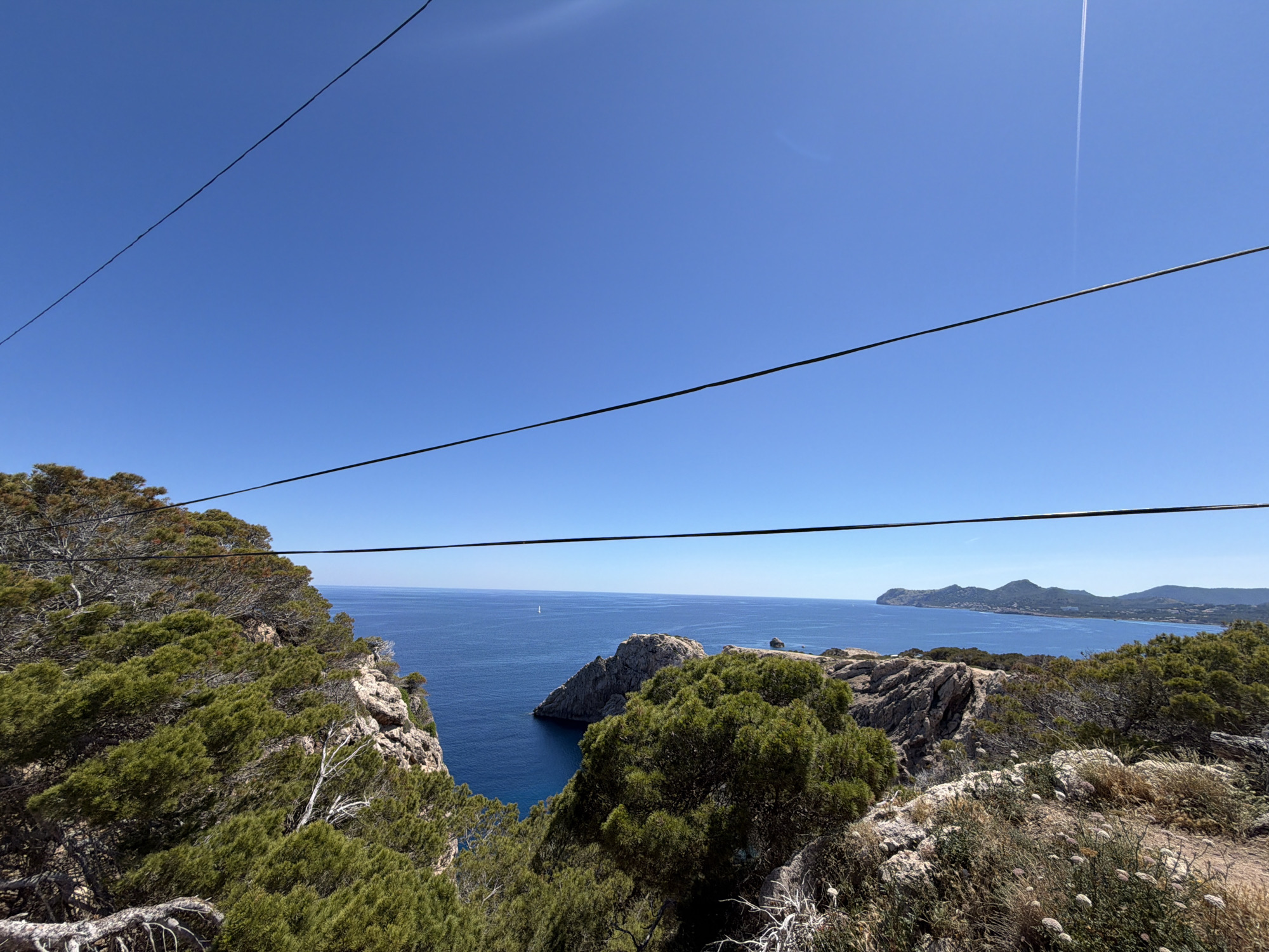 A bright, daytime, wide-angle shot shows a cliff-top view in Capdepera, Spain, overlooking the Mediterranean Sea. The azure sky is mostly clear, with a faint contrail from a high-altitude airplane. Two parallel electrical or communication wires bisect the upper part of the frame.
The viewpoint is heavily vegetated with low, scrubby trees and bushes, characteristic of the Mediterranean landscape. Exposed rock formations are scattered among the greenery.
The sea appears calm and stretches to the horizon. In the distance, across the water, is a headland of rolling hills, part of the Capdepera coastline. A small sailboat is visible in the sea.