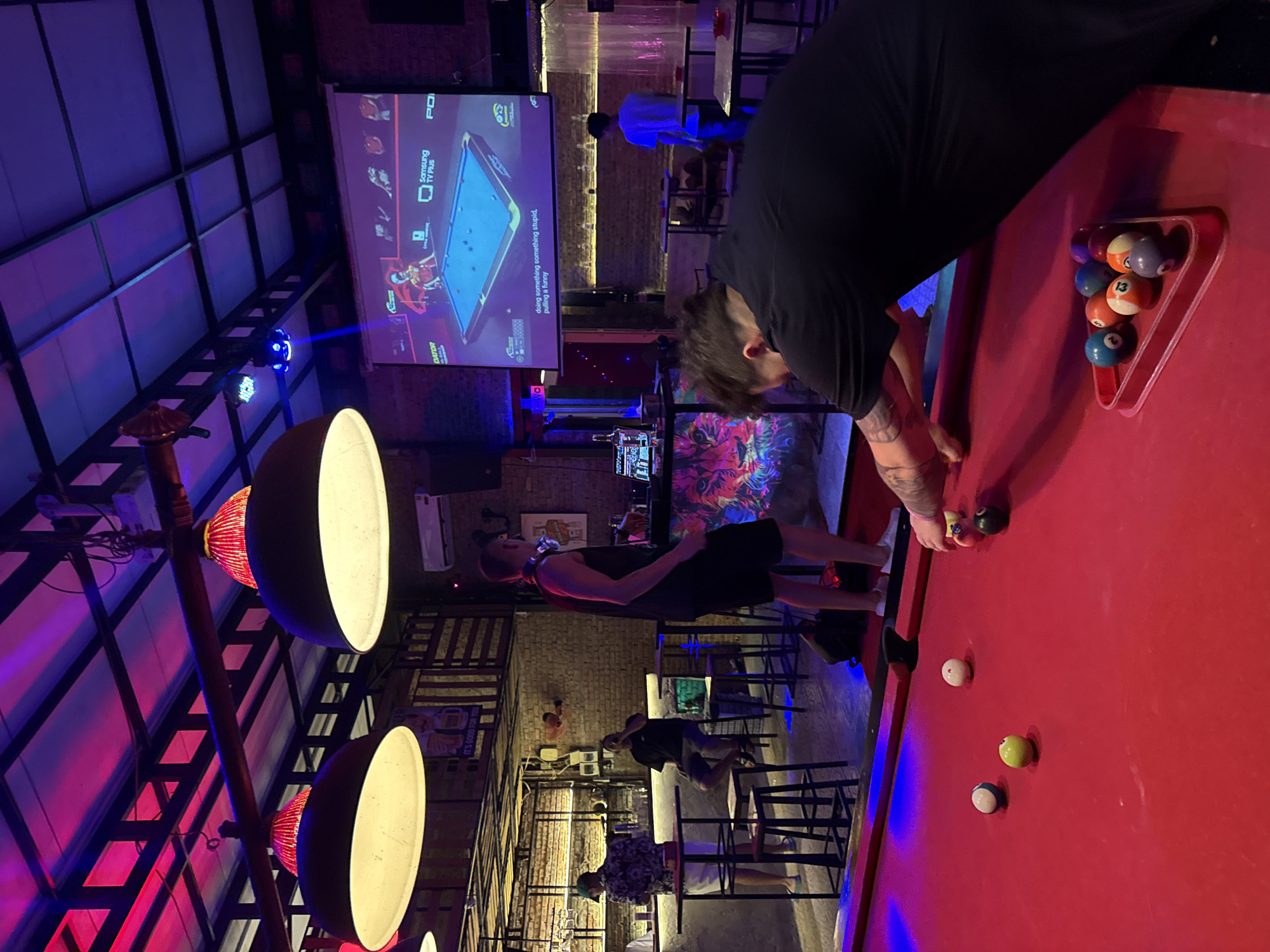 This image captures a vibrant indoor entertainment venue, likely a bar or lounge in Đà Nẵng, Vietnam, at night. The scene is illuminated by striking blue and purple ambient lighting, contrasting with the warm glow from large, bowl-shaped pendant lights and the bright red surface of a pool table in the foreground.
The primary activity centers around a red-felt pool table where a person wearing a black shirt with visible arm tattoos is leaning over to take a shot, holding a cue stick. Several pool balls are scattered on the table, including a white cue ball, a yellow ball, a blue and white striped ball, and a black 8-ball. A red triangular rack on the side of the table holds various numbered pool balls, including balls marked '6', '13', and '10'. Another individual, dressed in a black tank top and shorts, stands near the table, seemingly observing or waiting to play.
In the background, a large projector screen displays a pool game simulation or instructional content, featuring the text "Samsung TV Plus" and fragments like "PO" and "DATOR," along with the humorous phrase "doing something something stupid, pulling a funny." Below the screen, another person in a light blue shirt is partially visible, while further back, several patrons are seated or standing at tables and stools, socializing. One person wears a distinctive floral shirt.
The venue features an industrial-style ceiling with exposed beams and a track lighting system. A small disco ball or effect light emits blue beams across the room, contributing to the energetic atmosphere. Exposed brick walls add to the establishment's character, along with decorative elements featuring colorful, abstract patterns. The overall impression is one of a lively and relaxed setting for evening entertainment.