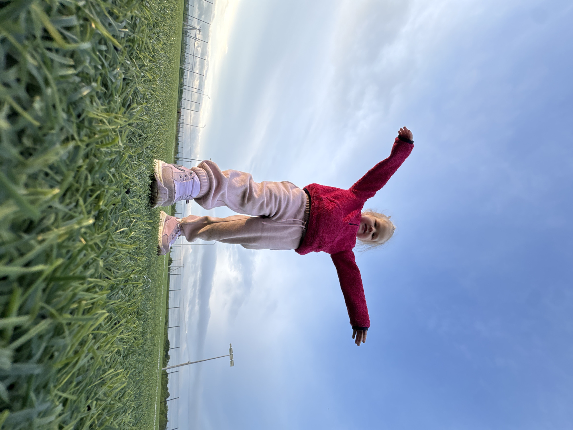 This low-angle photograph captures a young, fair-skinned child standing centrally on an artificial green turf field in Ферма, Russia. The child, clad in a fuchsia long-sleeved fleece top, light pink sweatpants, and light purple sneakers, extends both arms outwards, forming a horizontal line. Her gaze is directed towards the camera, and a subtle smile is present. The immediate foreground consists of sharply focused artificial green grass blades. In the midground and background, a chain-link fence delineates the field perimeter, behind which a white-framed soccer goal is visible on the left. Multiple utility poles and a tall floodlight tower with a rectangular array of lights are also present in the distance. The upper two-thirds of the image display a sky transitioning from clear blue on the right to partly cloudy with scattered white clouds on the left. The lighting suggests natural daylight, likely late afternoon or early evening.