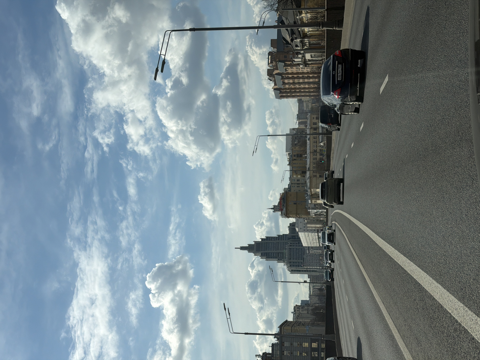 The image shows a roadway in Moscow, Russia, with cars traveling along it. The road has two lanes, separated by white lines. Streetlights line the side of the road. Buildings can be seen in the background on the right side of the image. The sky is cloudy, with patches of blue visible.
