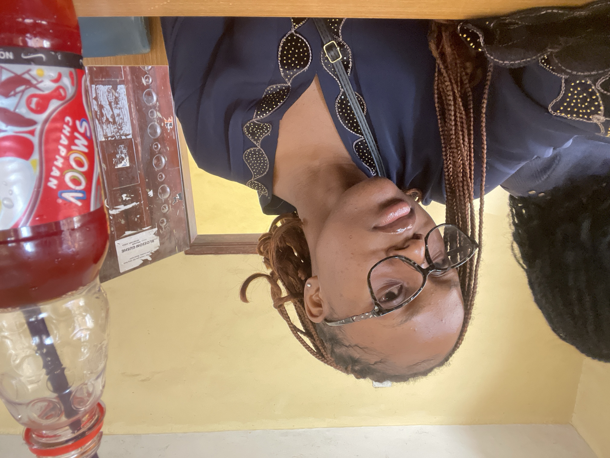 A medium shot shows a young Black woman with braided hair and glasses looking towards the camera. She is wearing a dark blue blouse with decorative gold bead embellishments around the neckline and sleeves, and a dark green strap across her chest. To her left, partially visible, is another person with dark, curly hair. In the foreground on the right is a clear plastic bottle filled with a red beverage, with a straw inserted. The background is a plain yellow wall. A door frame and part of a wooden cabinet are visible behind the woman. A small white sticker with text is affixed to the cabinet. The image appears to be taken indoors.