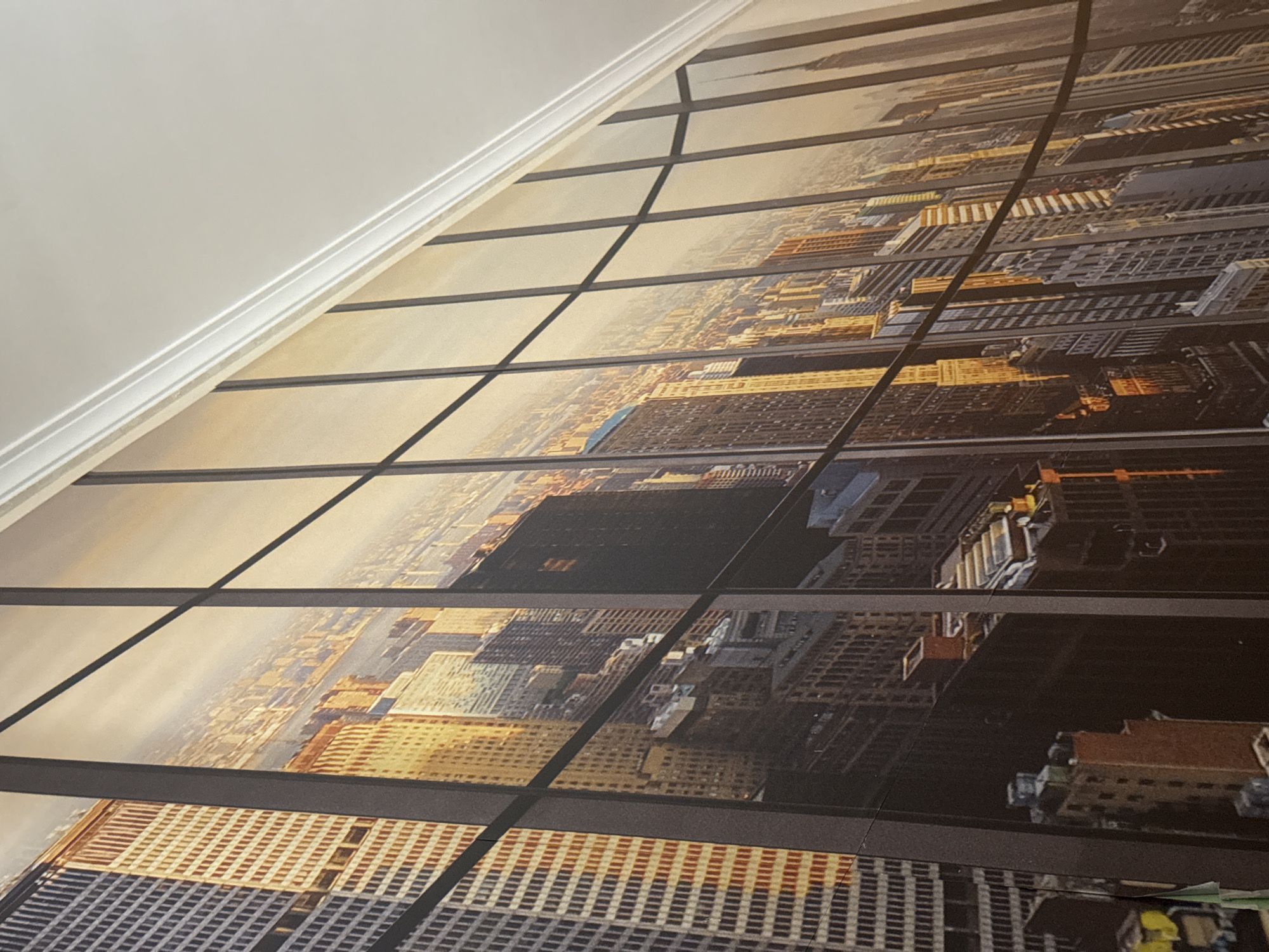 The image shows a perspective view of a cityscape, possibly New York, as seen through a window. The window is divided into multiple panes with dark frames that create a grid pattern. The city buildings vary in height and color, with a mix of beige, brown, gray, and white tones. The perspective suggests a high vantage point, looking down upon the city. There are no people visible in the image. The image does not appear to be in the city of Mirandela, Portugal, as the depicted architecture is characteristic of large North American cities.