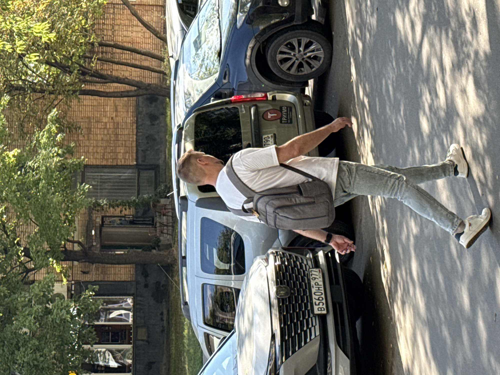The image captures a street scene in Moscow, Russia, under a bright, sunny sky casting dappled shadows from trees onto the pavement. A man, appearing to be in his late 30s or early 40s, walks away from the viewer towards the right. He is wearing a white t-shirt, light-colored jeans, and white sneakers. A grey backpack is slung over his shoulders.
In the background, several cars are parked. On the left, the front of a silver car with a distinctive grille and license plate "B560HP 977" is visible. To its right, a silver SUV is parked. Further to the right, a dark-colored SUV is parked, and a portion of another vehicle is visible on the far right. A building with a brick facade stands behind the cars, featuring windows and an open doorway. Trees with green foliage are prominent, with branches extending over the scene. The overall impression is a typical urban environment on a clear day.