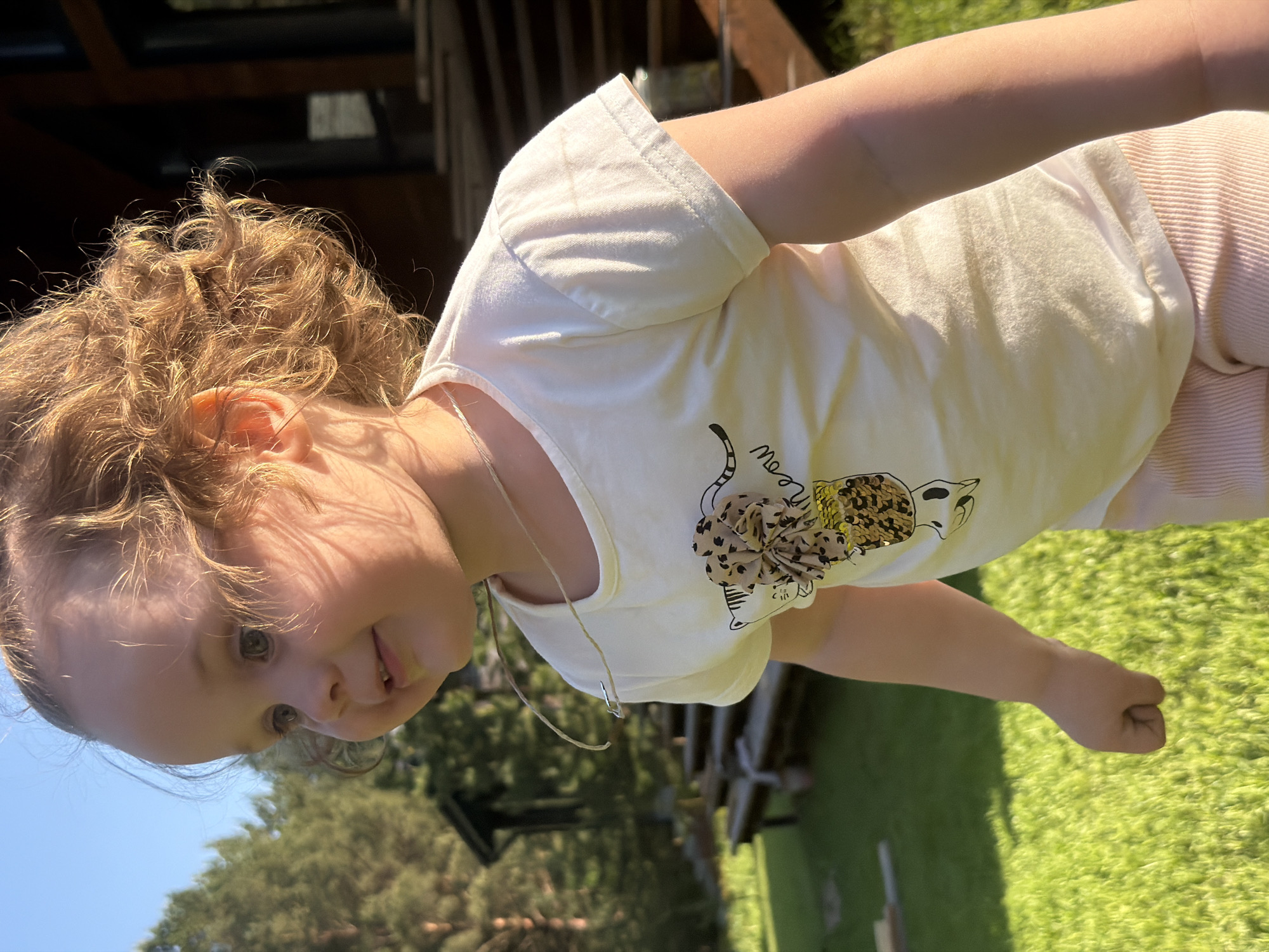 A young child with curly blonde hair and wearing a light-colored t-shirt with an animal design is captured in a close-up shot. The scene includes a grassy area with trees in the background, and part of a wooden structure is visible. The setting appears to be outdoors on a sunny day. The location is Kamyennoye, Russia.