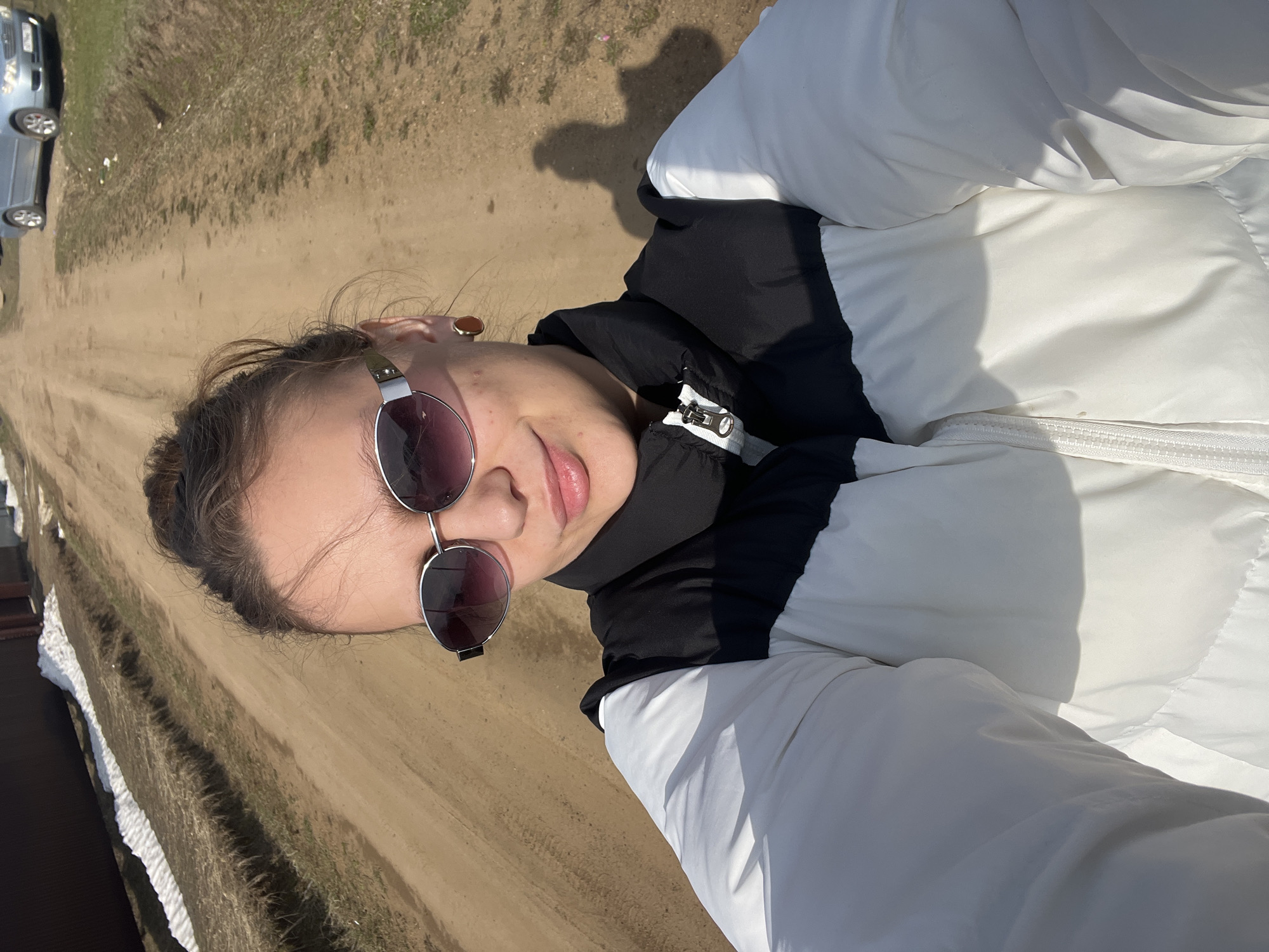 A woman in sunglasses and a white and black puffer jacket is centered in the image, likely a selfie. The background shows a dirt road and grassy areas typical of a rural or semi-rural setting. In the distance on the left side of the image, a parked silver car is visible. Given the location, this likely depicts a scene in or near Ольховка, Russia, although specific landmarks are not visible to confirm this.