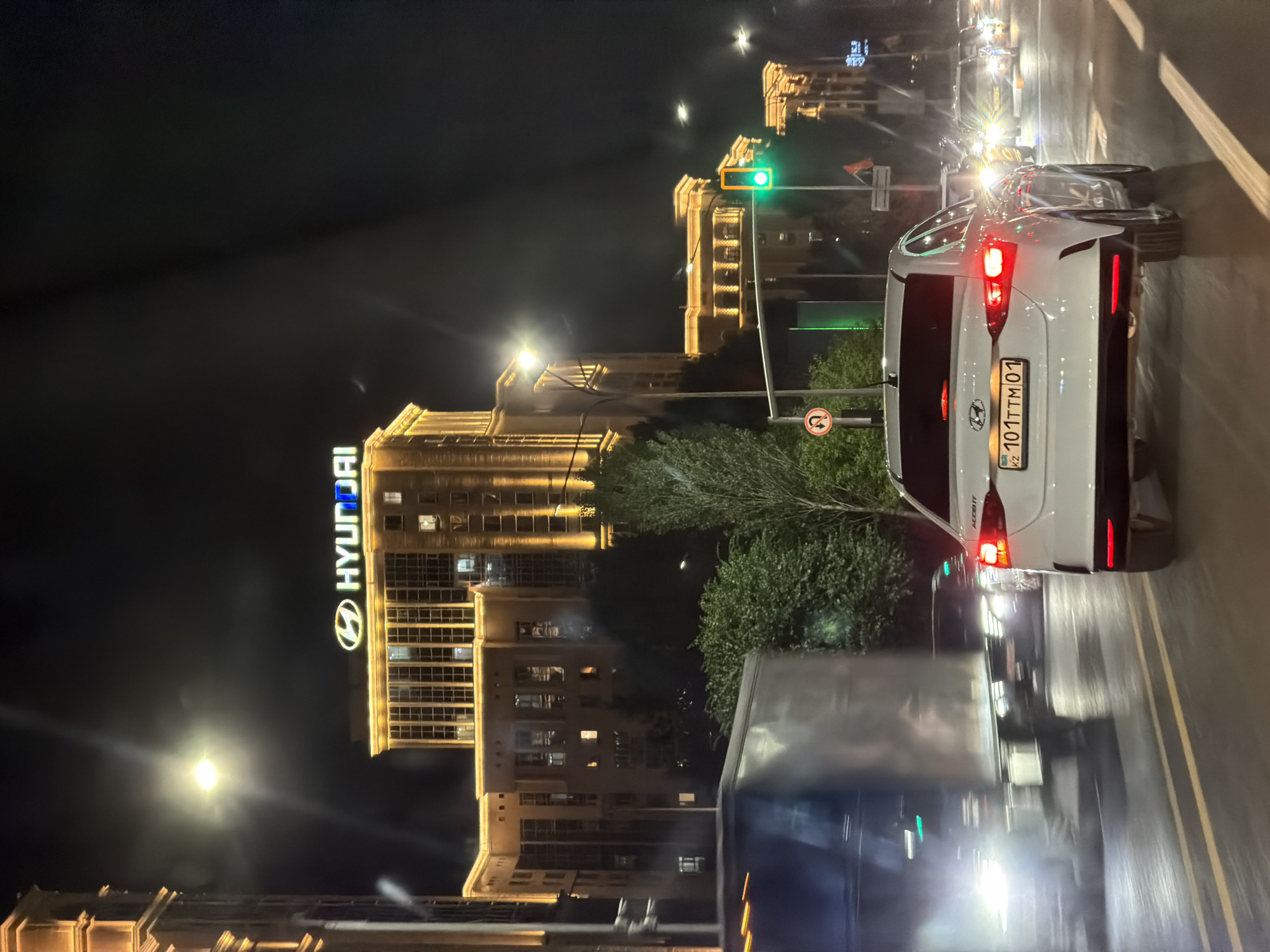 The image captures a night-time scene on a city street in Astana, Kazakhstan, likely a major avenue given the imposing illuminated architecture. A large, modern building with a prominent "HYUNDAI" sign in bright neon lettering dominates the upper background. The building is ornately lit with warm yellow lights, highlighting its architectural features. Multiple lanes of traffic are visible, with several cars in motion. In the foreground, a white Hyundai Accent is driving away from the camera, its brake lights illuminated. To the left, a large, silver truck is also moving, its headlights bright and creating a light mist. Traffic lights are visible, with one displaying a green light. Trees and vegetation are present along the side of the road, adding a touch of nature to the urban environment. The overall impression is that of a busy, well-lit city street at night.