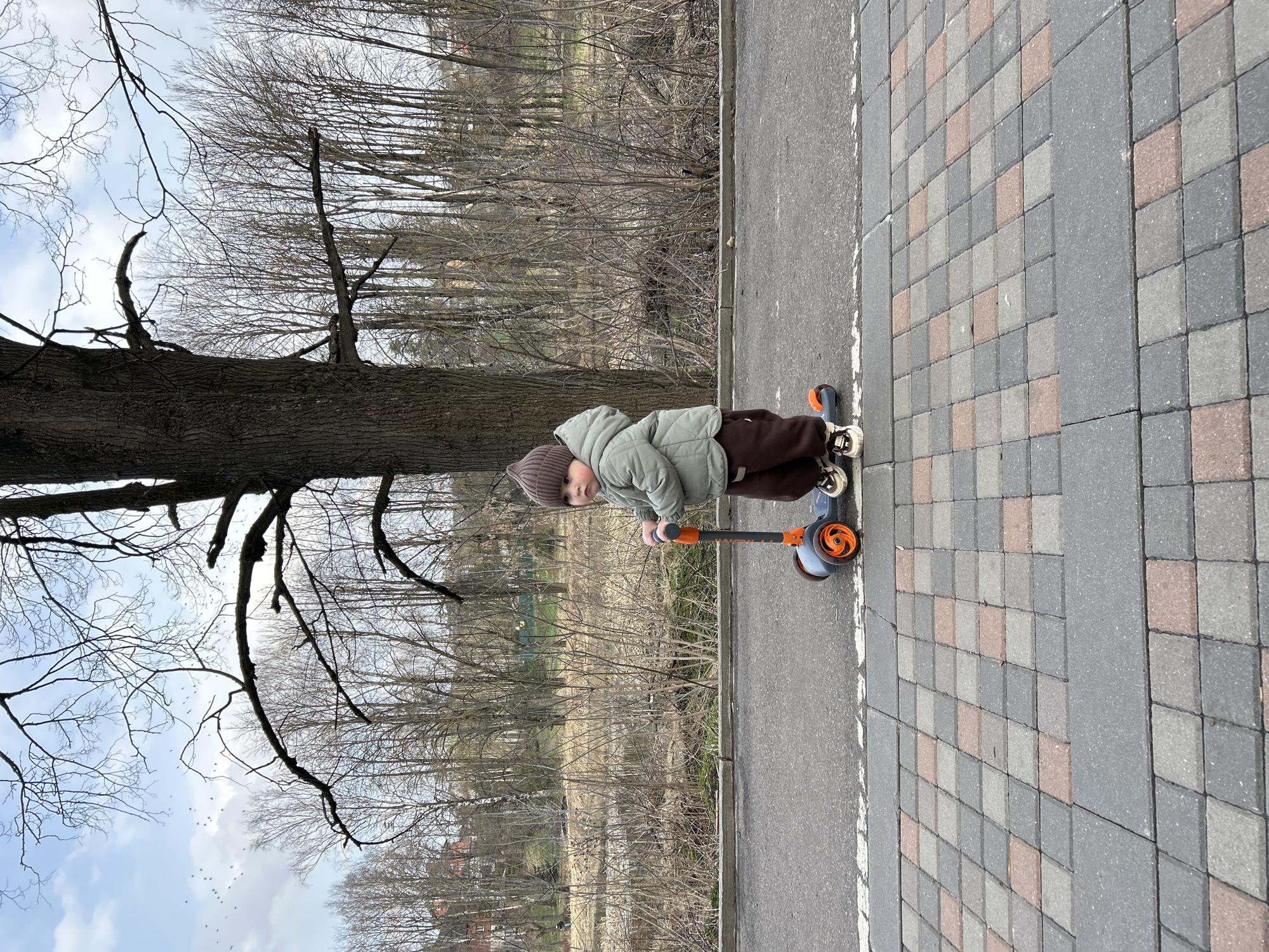 A child is on a scooter on a brick sidewalk next to a paved road in Klin, Russia, on a day with bare trees and a partly cloudy sky. The child wears a brown knit hat, light green jacket, and brown pants. The scooter is gray with orange accents. The sidewalk is constructed of gray and pinkish-brown bricks. The road surface is gray. Bare trees and underbrush line the other side of the road.
