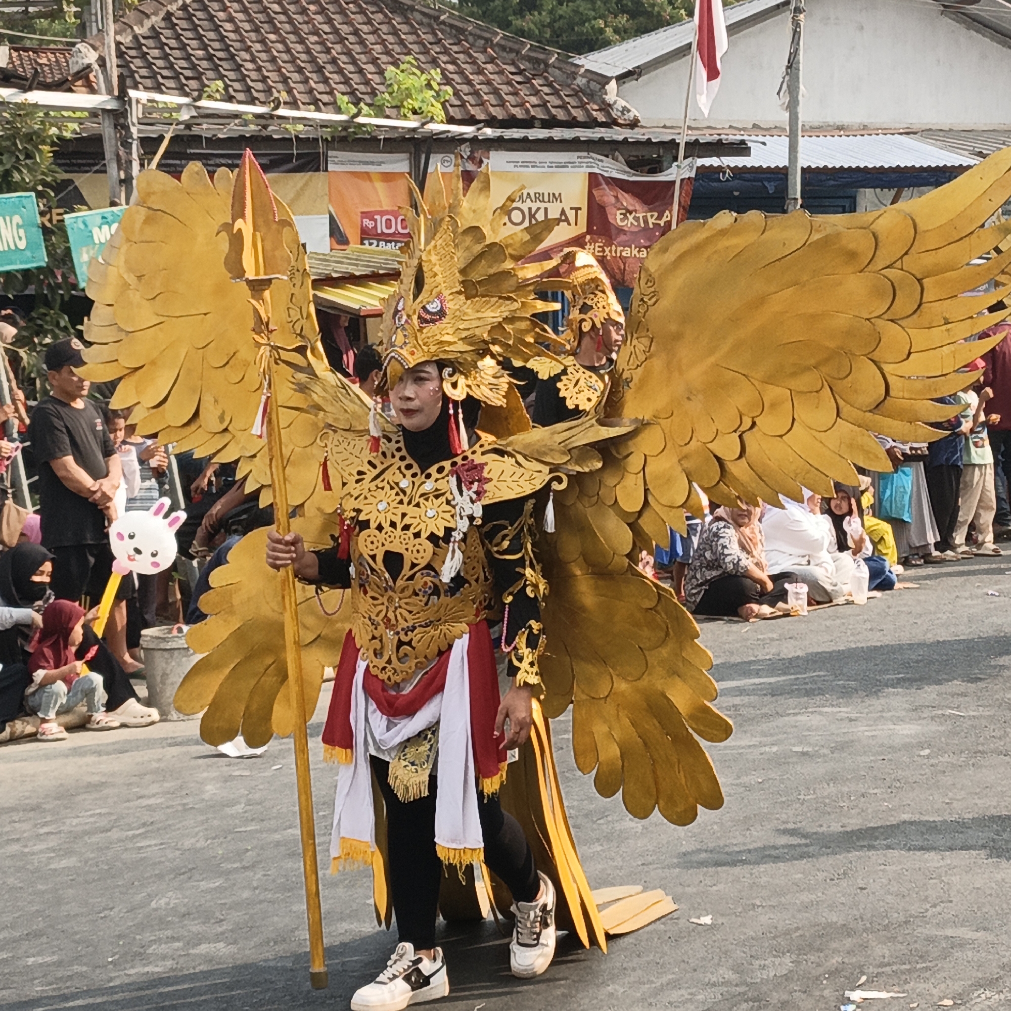 The image depicts a street parade in Randudongkal, Indonesia. A person in a large, elaborate golden costume resembling a mythical bird with prominent wings is walking down the street, holding a tall spear-like object. The costume features intricate gold detailing on the chest and headpiece, and is adorned with red and white fabric. They are wearing black leggings and white sneakers. In the background, a crowd of people are watching the parade, some seated on the ground and others standing. Various stalls and banners are visible behind the spectators, including one with Indonesian text and a picture of a cigarette pack. The event appears to be taking place outdoors on a paved street under a sunny sky.