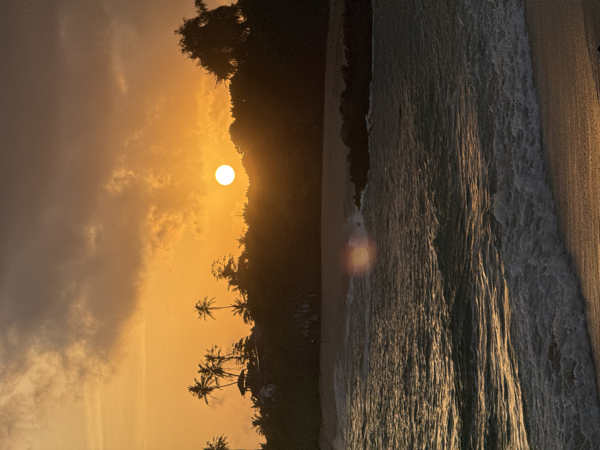 This is a vertical, sunset shot of a beach in Midigama, Sri Lanka. The sun is a bright white orb low in the sky, casting a warm orange and yellow glow across the scene. The sky is filled with soft, diffused clouds. In the foreground, palm trees are silhouetted against the vibrant sky. A gentle wave is breaking on the sandy shore, with white foam cresting. The water is dark and rippled, reflecting the warm light. The overall mood is peaceful and serene, capturing the beauty of a tropical sunset. There is no visible text in the image.