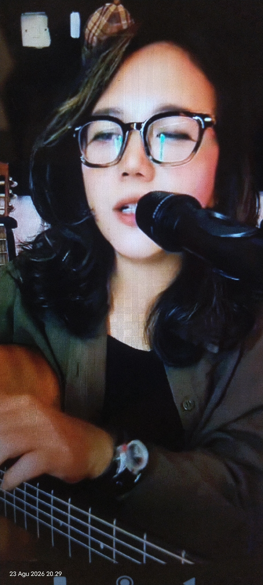 The image shows a close-up of a person wearing glasses and a plaid headband, holding a guitar and singing into a microphone. The person is wearing a dark shirt and a green jacket. The background is dark, with some blurry objects that could be part of a room. The bottom left of the image shows the date "23 Agu 2026 20.29". The scene does not provide any specific details about a location in the city of East Jakarta, Indonesia.