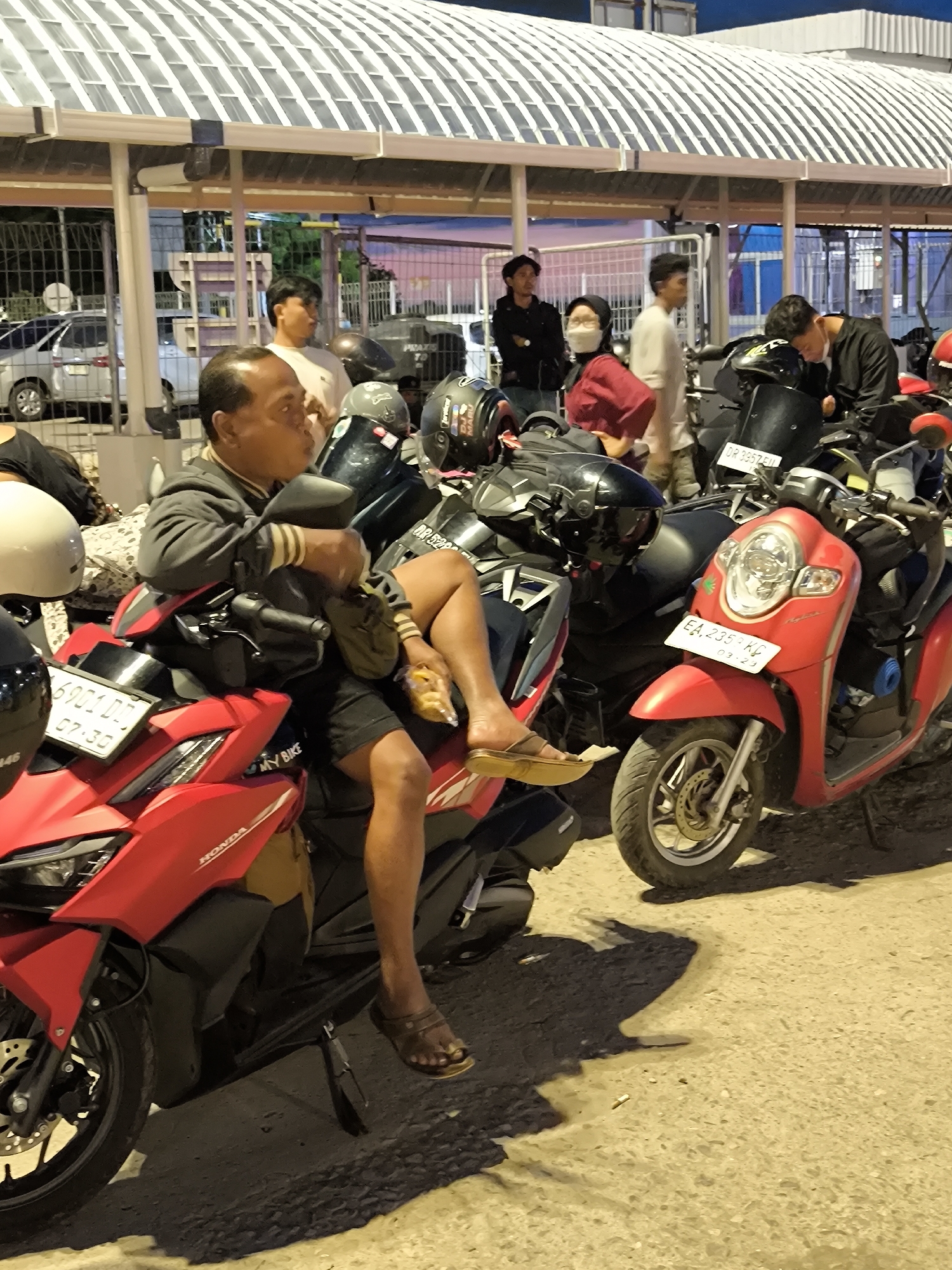 The image shows a man sitting on a red motorcycle in an outdoor parking area or street scene. The man is wearing a gray jacket, black shorts, and brown sandals, and is holding a small bag or snack in his hand. Several other motorcycles are parked nearby, including a red scooter and another larger bike. A group of people is standing and interacting in the background near a covered walkway or building structure with a white slatted roof. The sky is transitioning from dusk to night. License plates visible on the motorcycles read "6901 DD 07-30", "DR 3957 FIL", "DR 52", and "EA 2359 KC 03-23".