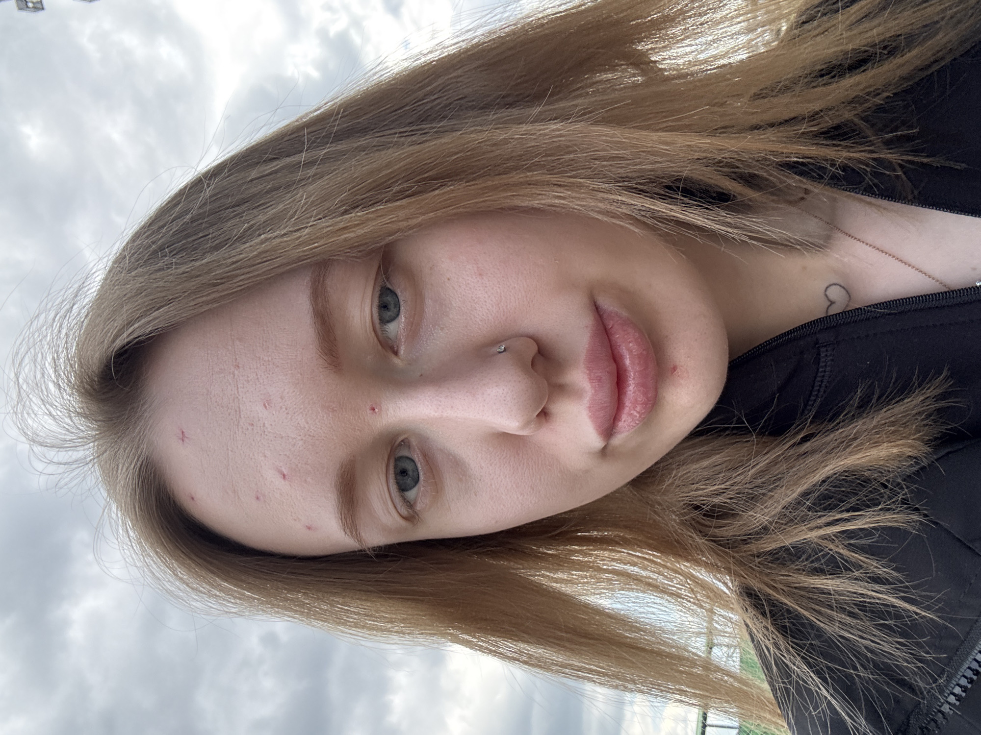 A close-up selfie shows a young woman with fair skin and long, light brown hair, angled looking directly at the camera. Her head is tilted, and the image is rotated approximately 90 degrees counter-clockwise, making the sky appear to her right. She has light blue or green eyes and a neutral expression with slightly parted lips. A small silver stud piercing is visible on her right nostril. Several small red blemishes are present on her forehead and left cheek.
She is wearing a dark, possibly black, zipped jacket or top, the zipper pull visible on the right side of the frame. A delicate, thin gold-colored necklace is around her neck. Below the necklace, a small, simple black outline of a heart is tattooed on her upper chest/collarbone area. The background consists of an overcast sky with scattered clouds, indicating an outdoor, daytime setting. No specific location cues or visible text are discernible in the image.