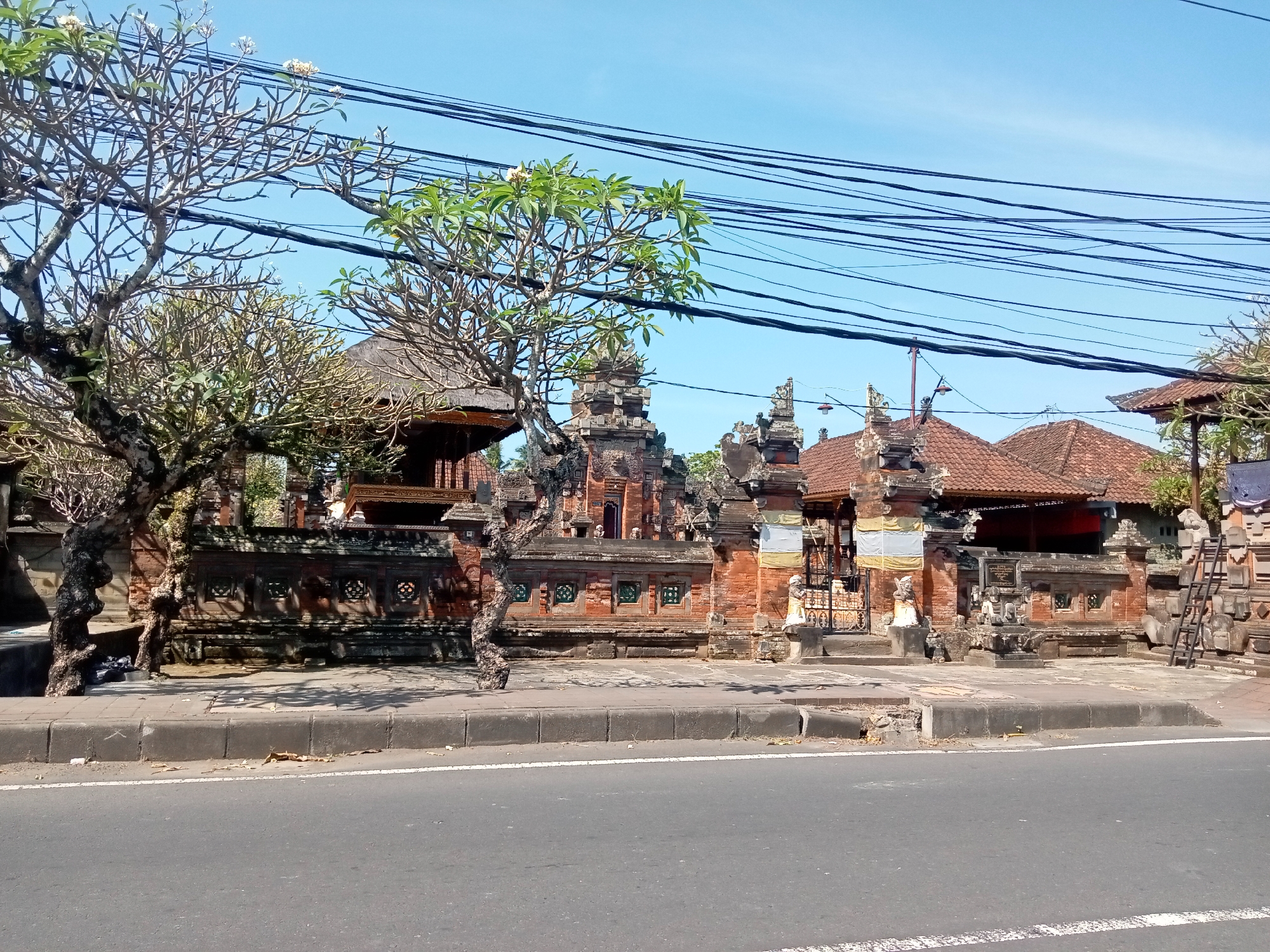 An outdoor, eye-level, full shot captures a street scene in front of a traditional Balinese temple. The temple, constructed from brick and stone, features ornate carvings, tiled roofs, and several gates. Two figures, likely statues or representations of deities, stand guard at one of the gates. A large, leafless tree with a gnarled trunk is prominent on the left, its branches reaching towards the sky, interspersed with power lines. The street in the foreground is asphalt, with a concrete curb and sidewalk. The sky is clear and blue.