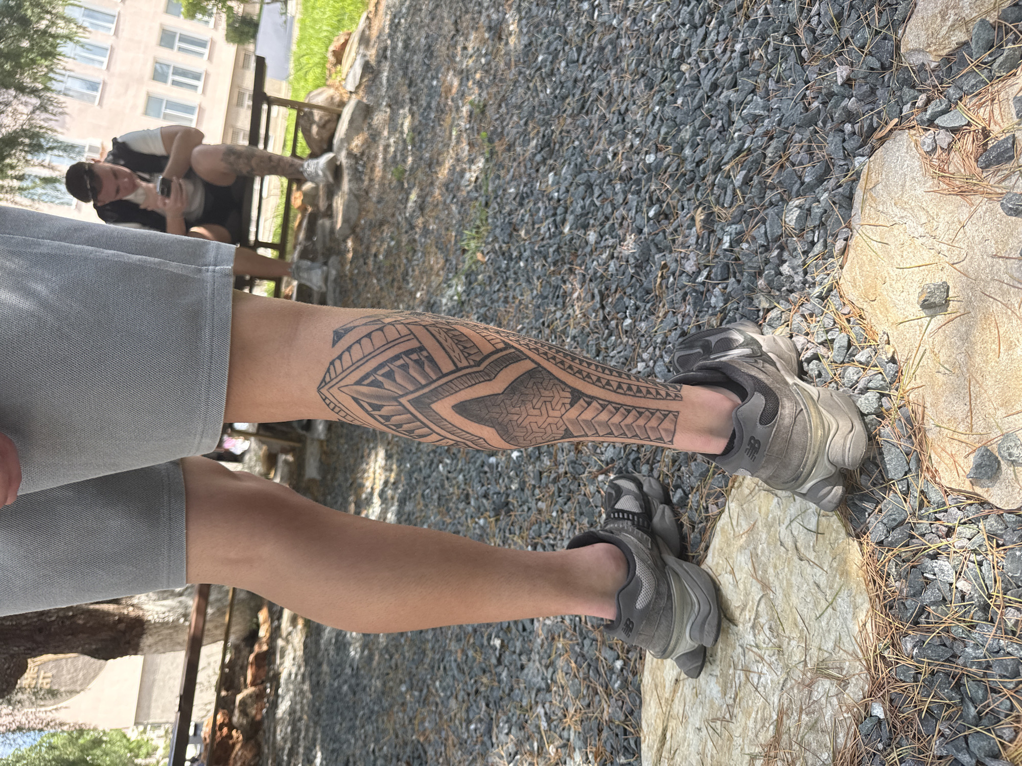 A person in grey shorts stands on a grey gravel surface with a large leg tattoo on their right calf. The tattoo is black and features geometric patterns. The person wears grey New Balance athletic shoes. A large, light-colored stone is visible to the right of the person. In the background, a man sits on a bench, engaged with his phone. A building is seen behind the bench. It is likely the image was taken outdoors in a public area such as a park, most likely in the city of Elhovo, Bulgaria.