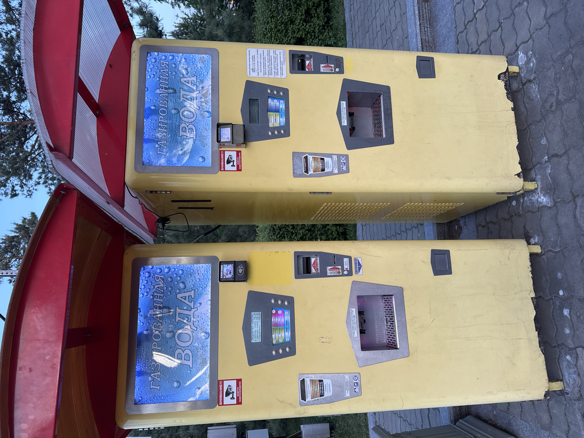 This outdoor shot, likely taken in Khabarovsk, Russia, shows two yellow soda vending machines. The machines feature blue screens with text in Russian. Each machine has a payment slot, a screen with options, and a dispenser area. The machine on the left is partially covered by a red and white awning. The right edges of the machines are damaged. The machines are located on a paved surface near some trees.