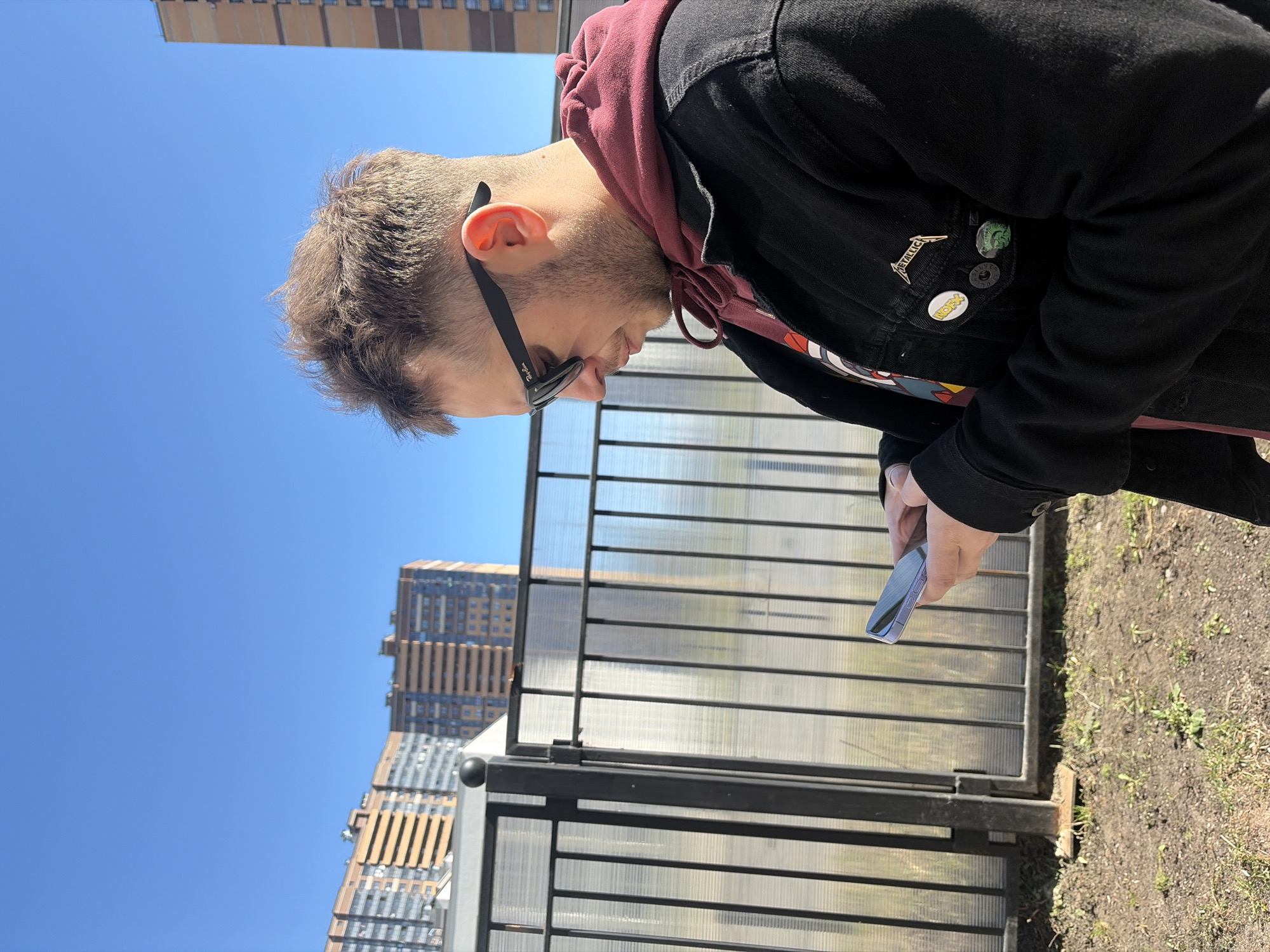 A person stands in what appears to be Saint Petersburg, Russia, based on the architectural style of the high-rise buildings in the background and the overall environment. The individual is wearing sunglasses, a maroon-colored hoodie, and a black denim jacket with various pins on it. They are looking down at a mobile phone held in their right hand. The scene is outdoors on a sunny day, with a blue sky overhead. A black metal fence with a semi-transparent barrier is located near the person, possibly separating a pedestrian area from a garden or green space. The ground shows some grass and bare earth.