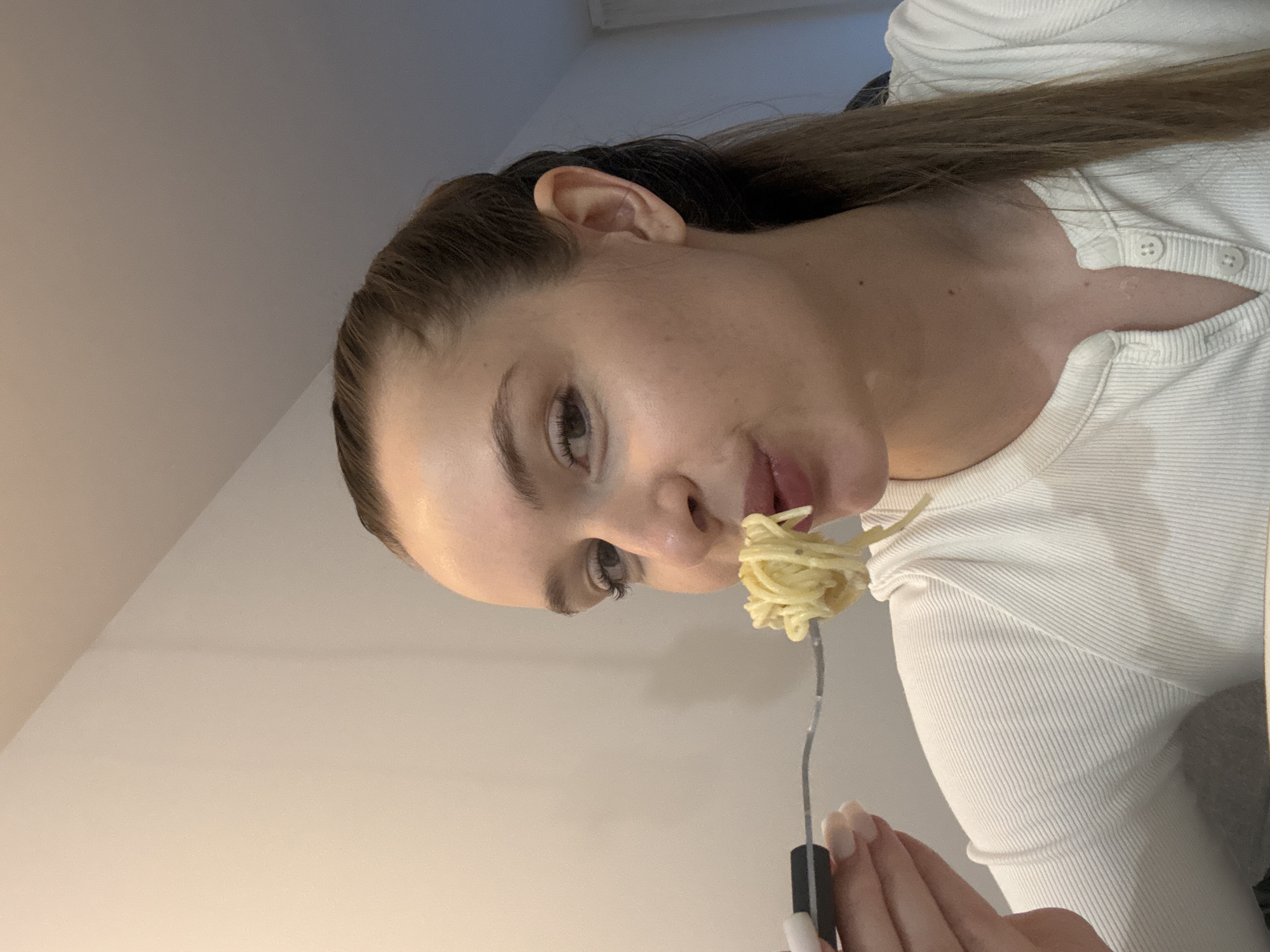 A young woman with brown hair, wearing a white, ribbed shirt, is eating spaghetti from a fork. She is indoors, likely in a home or apartment. The background is a plain, light-colored wall and ceiling. The location is not discernible from the image. The presence of Budva, Montenegro, cannot be determined from this photo.