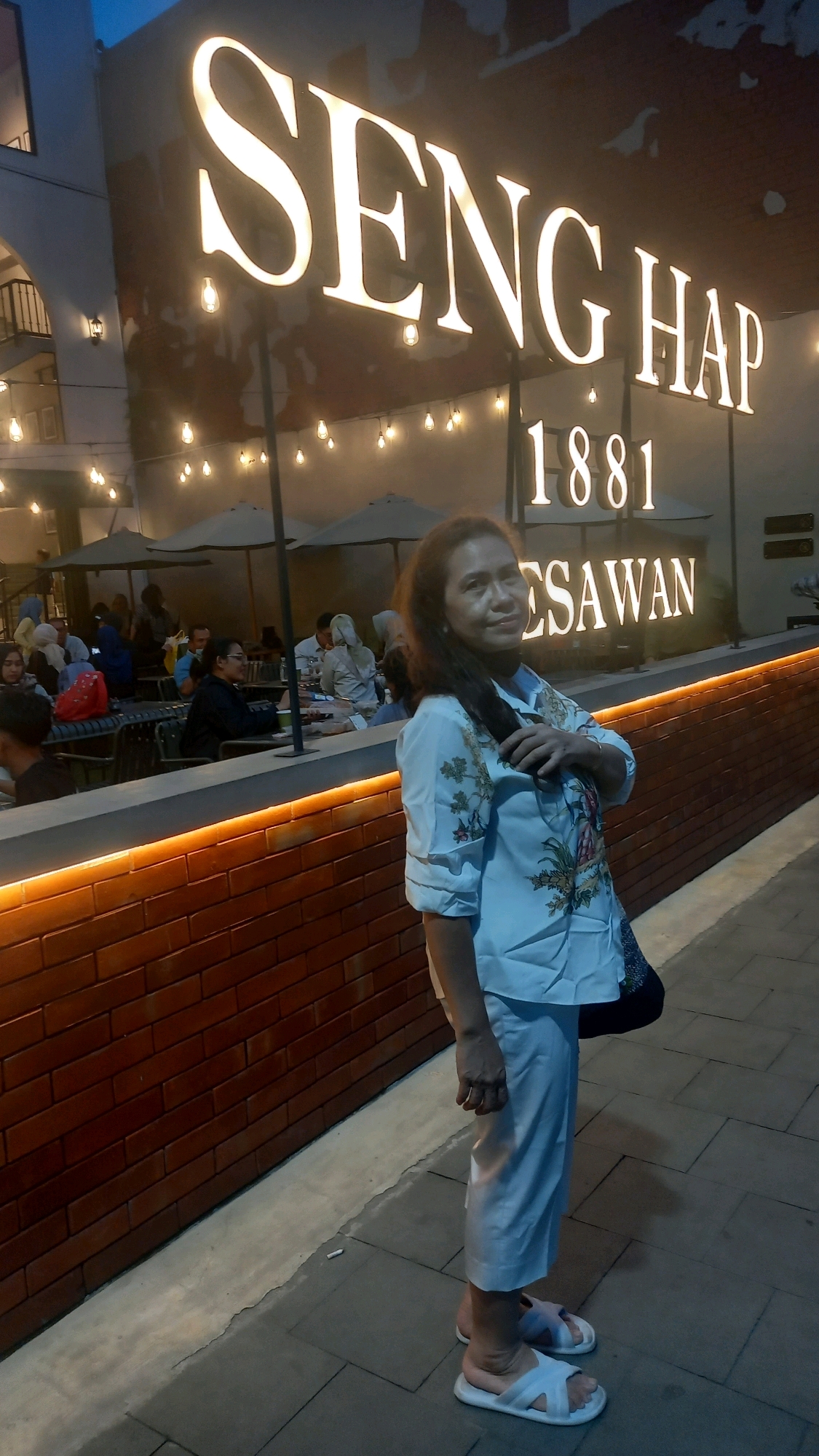 A middle-aged Asian woman stands in front of a building with a large illuminated sign that reads "SENG HAP 1881 KESAWAN" in City of Medan, Indonesia. She is wearing a light-colored long-sleeved shirt with a floral print and matching light-colored pants, along with white slide sandals. Her right hand is raised and resting on her chest. In the background, patrons are seated at tables under umbrellas in an outdoor seating area. The building facade features exposed brick with an orange illuminated strip. The scene appears to be a restaurant or cafe.