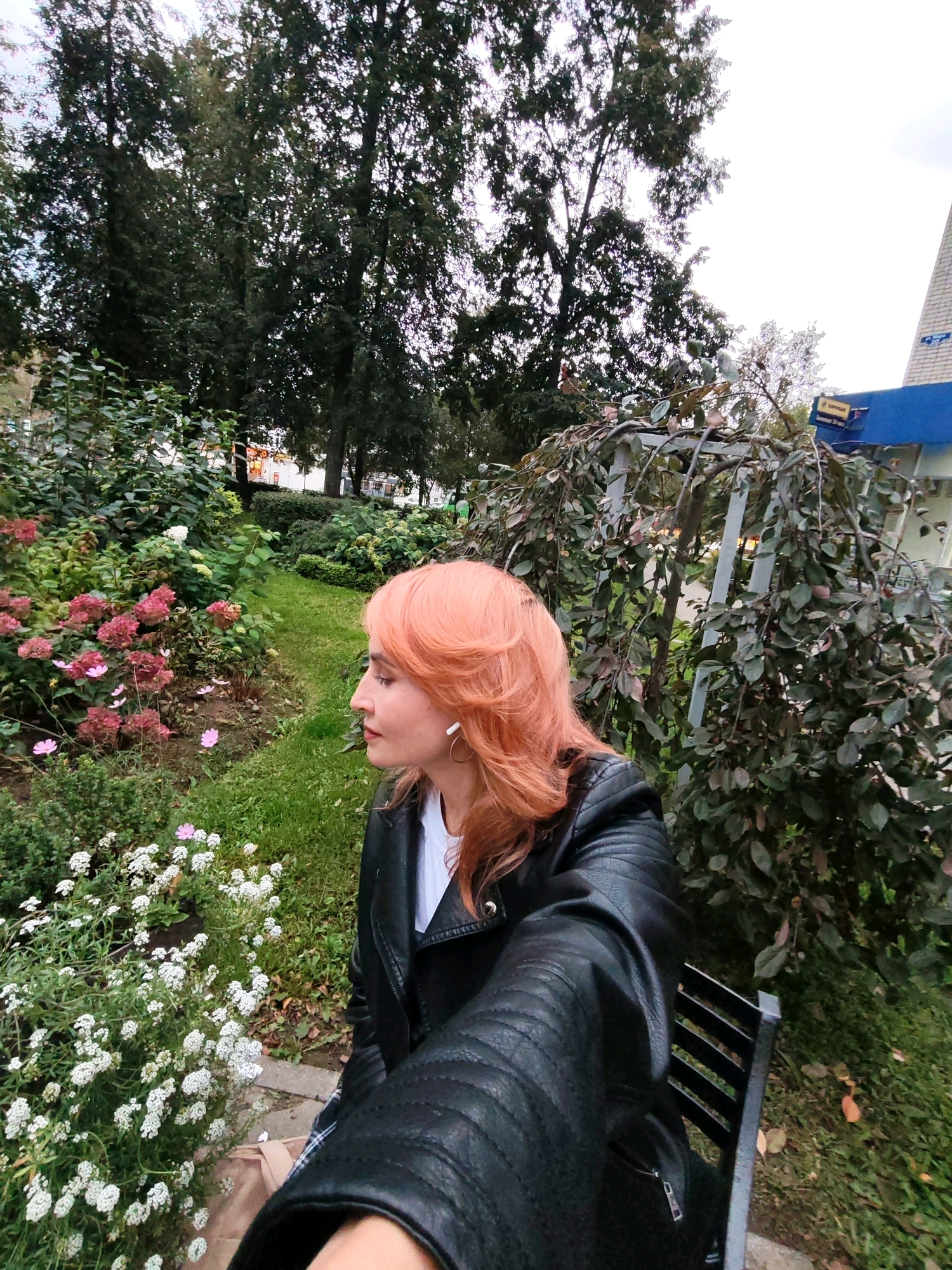 An individual with vibrant, coral-colored hair is seated on a dark-colored bench in a park-like setting. They are wearing a black leather jacket over a white t-shirt and have white earbuds in their ears. The person is looking to their left, with their head tilted slightly.
The background features lush greenery, including a variety of flowering plants and trees. To the left of the individual, there are clusters of pink and white flowers. To the right, a large bush with dark leaves and a metal support structure is visible. In the distance, a blue building with signs is partially obscured by the foliage. The ground is covered with grass and scattered autumn leaves. The overall environment suggests an outdoor public space, likely a park or garden within the city of Chaikovsky, Russia, given the provided context.