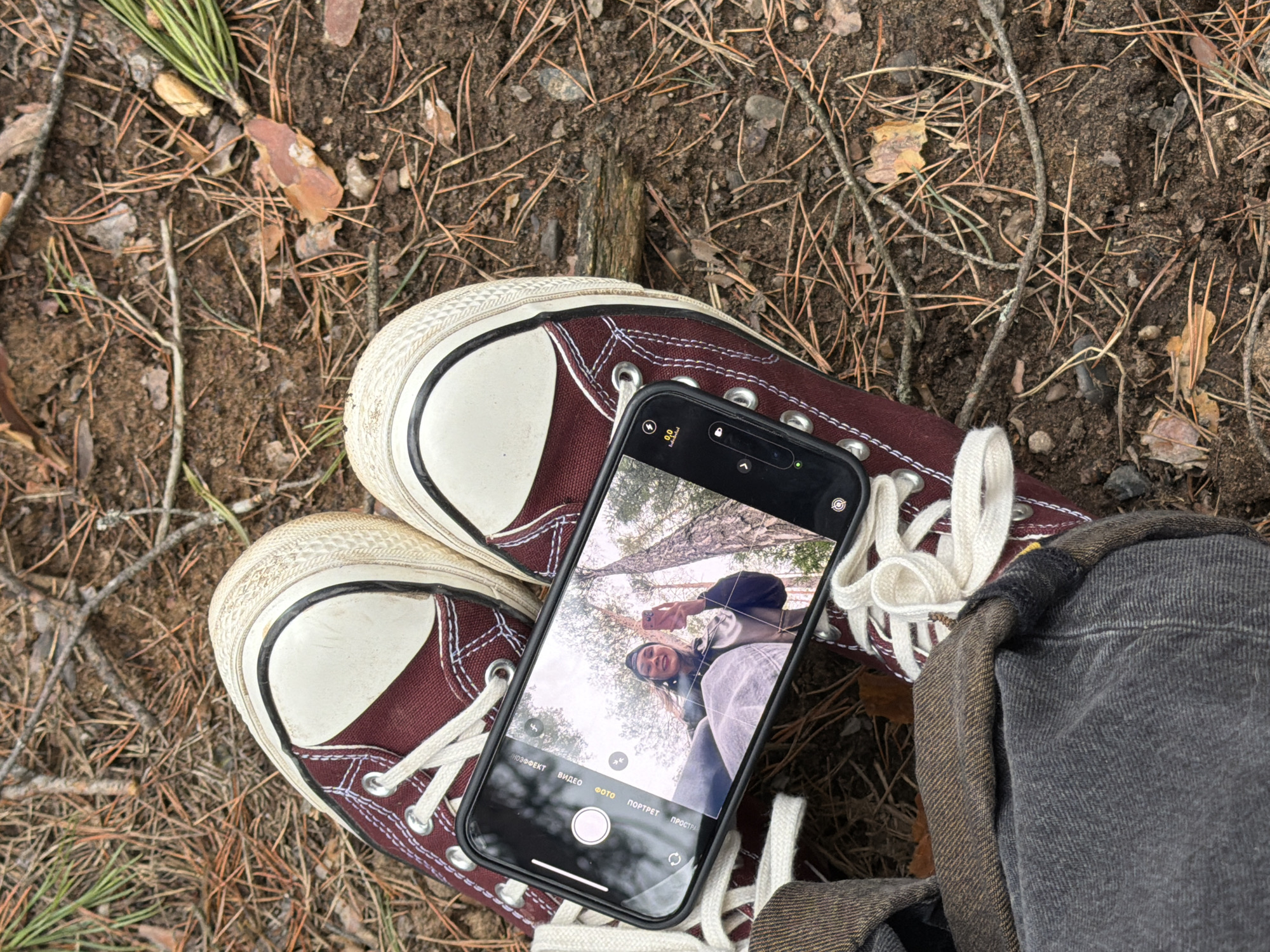 The image offers a top-down perspective of a forest floor, likely in Chaikovsky, Russia. The ground is composed of dark soil densely covered with fallen pine needles, small branches, and scattered dried leaves, suggesting a natural outdoor environment, possibly during autumn.
A pair of maroon high-top canvas sneakers with white laces and toe caps rests on this earthy surface. A dark-colored smartphone, identifiable as an iPhone by its interface and notch, is placed on one of the sneakers. Its screen displays a live camera feed or a reflection of a young woman. She is smiling broadly and looking upwards, holding another phone to capture a selfie or be filmed. She wears a dark hat or beanie and a light-colored top. The background within the phone screen shows tall trees against a bright, clear sky, confirming the outdoor, daytime setting.
To the right, a segment of charcoal-gray fabric, possibly from a jacket or trousers, is partially visible. The smartphone's camera app is active, with Russian text indicating various modes: "ВИДЕО" (VIDEO), "ФОТО" (PHOTO) which is currently selected, and "ПОРТРЕТ" (PORTRAIT). Other small camera icons and settings, such as "0.0" and a lock symbol, are also discernible. The scene captures a casual, happy moment of smartphone photography amidst nature.