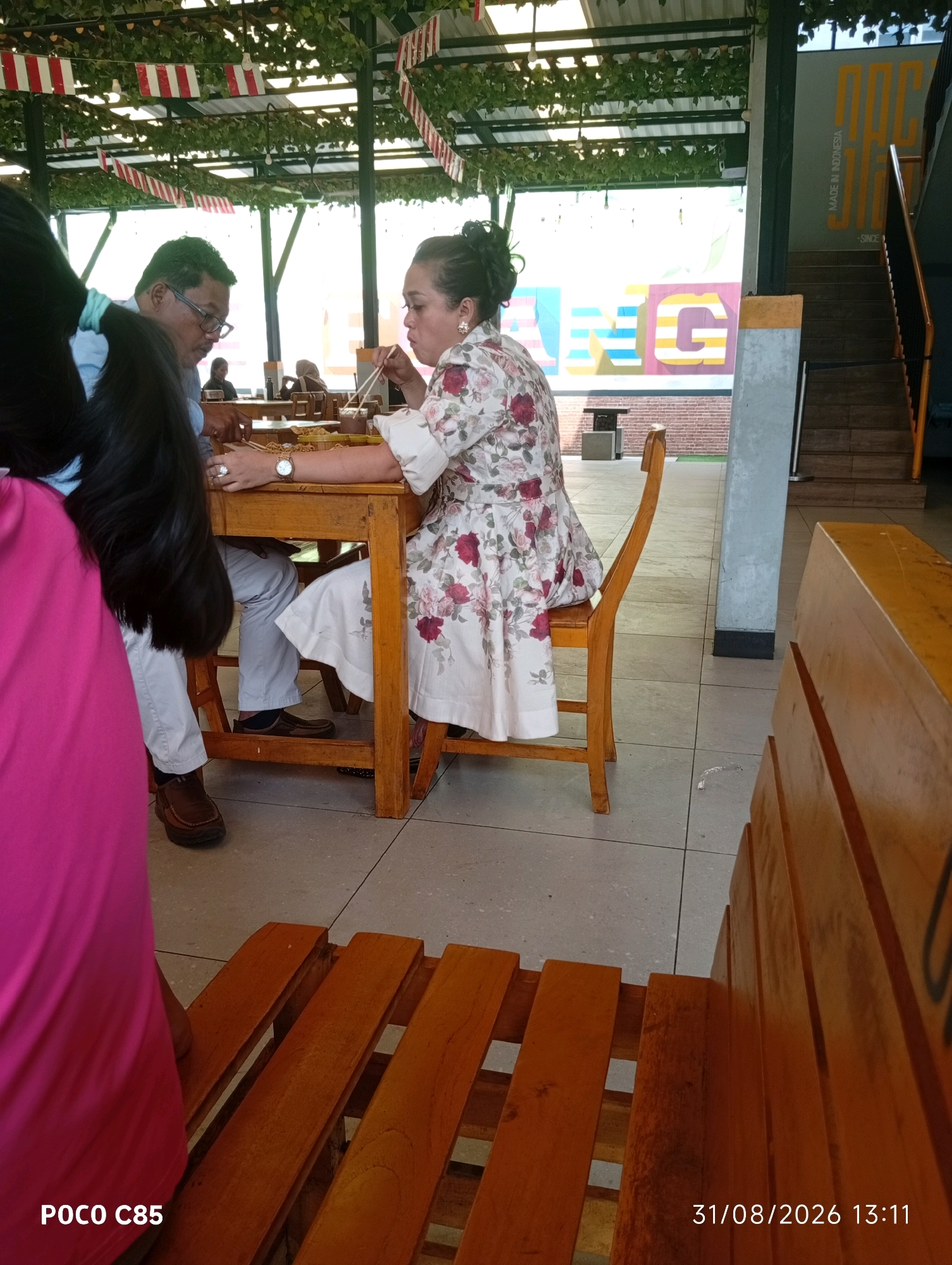 The image depicts an indoor dining area, likely a restaurant, with a contemporary design. The setting features wooden furniture, including tables and chairs, arranged on a tiled floor. Overhead, a decorative canopy of greenery and festive red and white banners creates a vibrant ambiance. A prominent wall mural with colorful letters and the text "MADE IN INDONESIA" suggests a connection to Indonesian culture and manufacturing.
In the foreground, a woman in a floral print dress is seated at a table, actively eating with chopsticks. To her left, a man in glasses and a blue shirt is also seated at the table, looking towards the woman. Partially visible in the left of the frame is the back of another person wearing a bright pink garment.
The overall scene conveys a casual dining experience within a visually engaging and culturally relevant environment. The presence of the Indonesian flag-like banners could indicate a patriotic theme or a celebration. The location in South Tangerang, Indonesia, is suggested by the text on the mural.