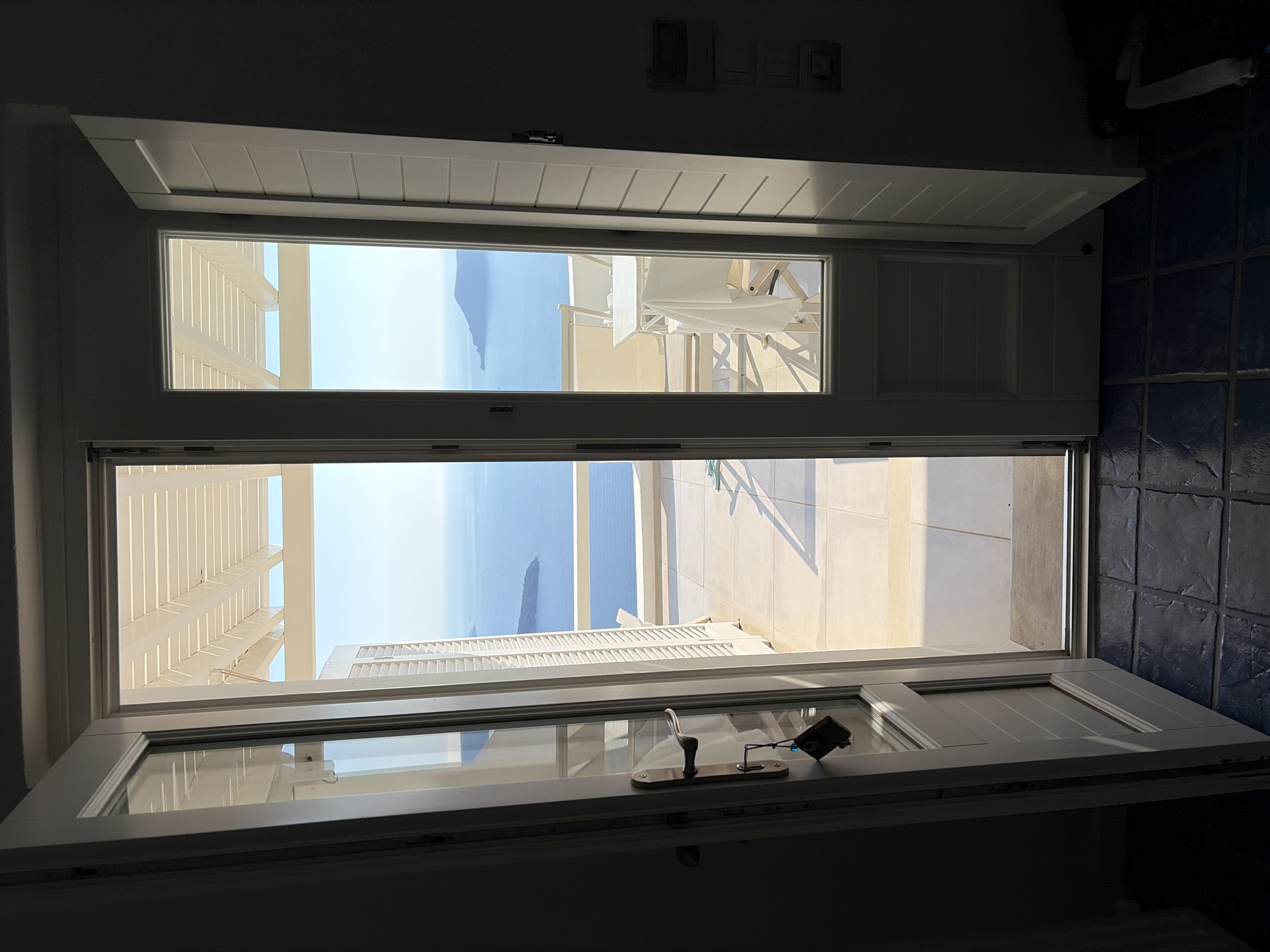 The image shows an open doorway with a view of the Aegean Sea and a balcony in Thira Municipal Unit, Greece. The doorway is framed by white doors, one of which is open. The balcony is furnished with a table and chairs, and a white tablecloth hangs from the table. The sea is a deep blue, and several islands are visible in the distance. The sky is clear and bright. The foreground shows a tiled floor.