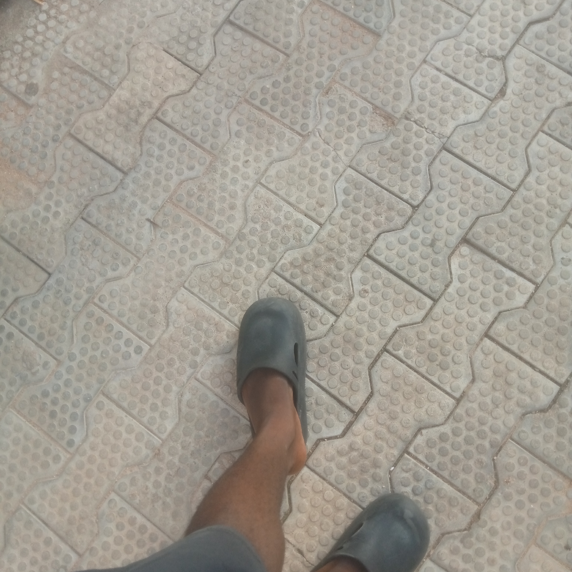 A top-down photographic view depicts the lower legs and feet of an individual standing on a textured, paved surface in the city of Unknown, Unknown. The pavement consists of light grey interlocking concrete pavers, each featuring a pattern of uniformly spaced, small raised circular dots. The pavers are characterized by an H-shape or dog-bone design, with narrow gaps between them showing traces of sand or dirt. The overall surface appears dusty and shows minor wear.
The individual's bare lower legs are visible, displaying a medium to dark brown skin tone and some body hair. Both feet are clad in dark grey, closed-toe, slip-on shoes, resembling clogs or sandals, made of what appears to be rubber or synthetic material. The shoe on the left foot, positioned more centrally, exhibits a small, elongated oval cutout on its upper side. A sliver of a dark grey garment, possibly shorts or trousers, is visible at the very bottom left edge of the frame. The person appears to be in a static position or in the process of walking, with the left leg slightly more prominent than the partially visible right leg.