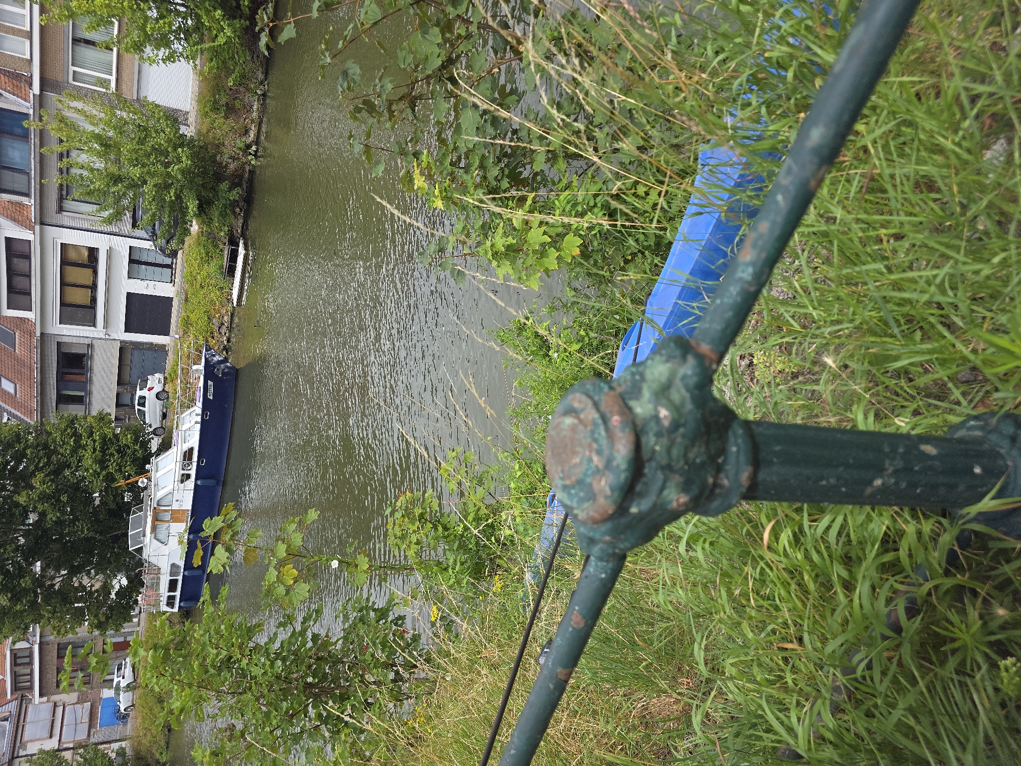 The image shows a canal in the city of Ghent, Belgium. A white and blue boat is docked on the left side of the canal, with several people visible on its deck. Residential buildings with red-tiled roofs line the opposite bank of the canal. Lush green vegetation, including trees and tall grass, grows along the canal banks. A rusty green metal railing is in the foreground, partially obscuring a bright blue object on the grassy bank. The water of the canal is calm with ripples on its surface.