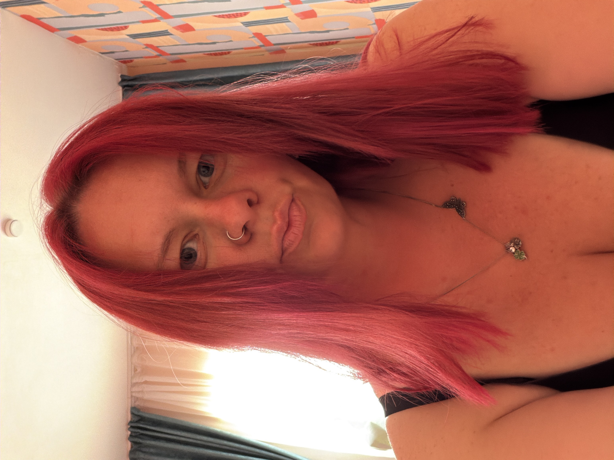 A woman with red hair is facing the camera, with a silver nose ring and a clover-shaped necklace. She is wearing a black top. The background includes a colorful patterned headboard and a blue curtain. The location cannot be determined based on the visual information provided.