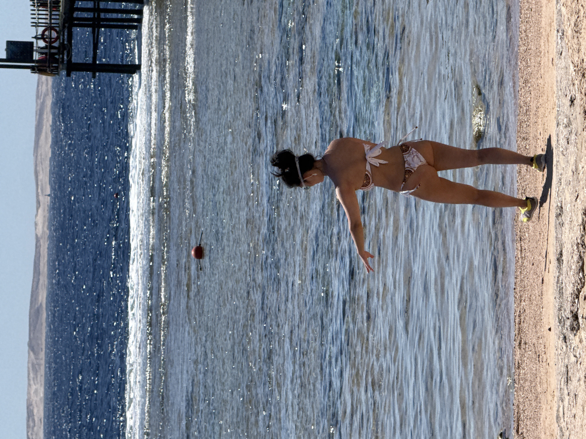 A woman in a patterned bikini stands on a sandy beach, facing the sea. She is wearing a headband and sunglasses, and her hair is pulled back. Her left arm is extended, and her hand is open, as if reaching out. Her right leg is slightly forward, and she is wearing yellow and grey athletic shoes. The sea is calm, with small waves breaking near the shore. In the distance, a red buoy is visible in the water, and a pier extends from the right side of the frame. The sky is clear and blue, with a hazy shoreline in the far distance. The image appears to have been taken in Sharm El Sheikh, Egypt, a popular tourist destination known for its beaches and diving sites.